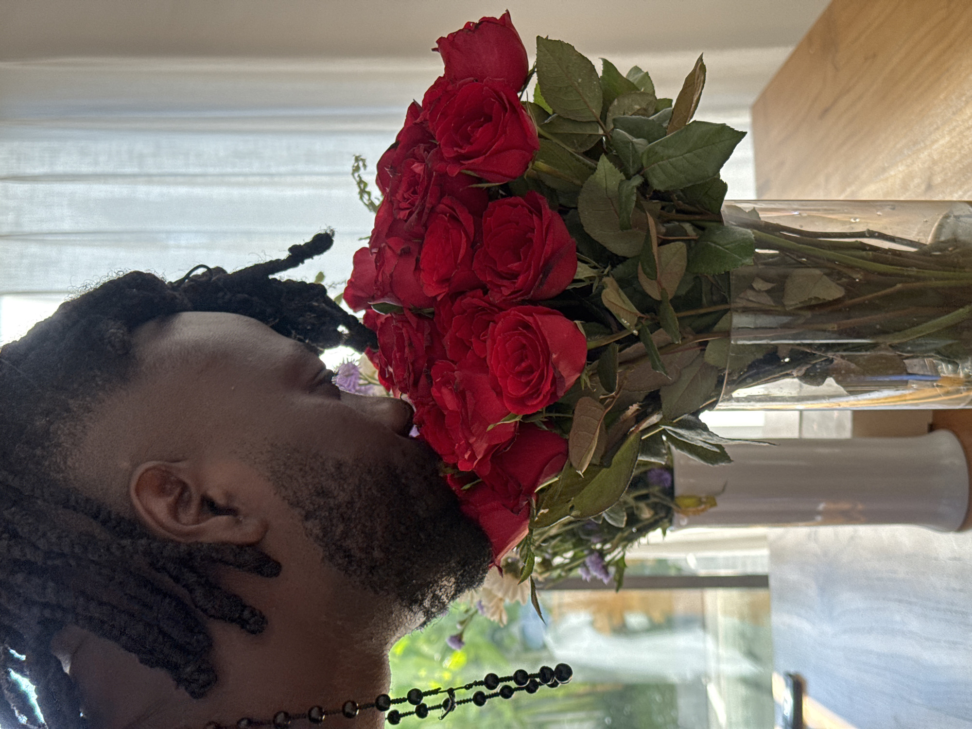 A close-up, medium shot shows a person with dark dreadlocks smelling a bouquet of red roses in a glass vase. The person is partially visible from the shoulders up, with their face turned towards the flowers, obscuring their eyes. They are wearing a necklace with dark beads. The bouquet is large, with many red roses and some smaller purple and white flowers. The arrangement is placed on a wooden table. In the background, there is a window with sheer white curtains and a view of greenery outside. The overall lighting suggests it is daytime. The image is captured from a slightly low angle.