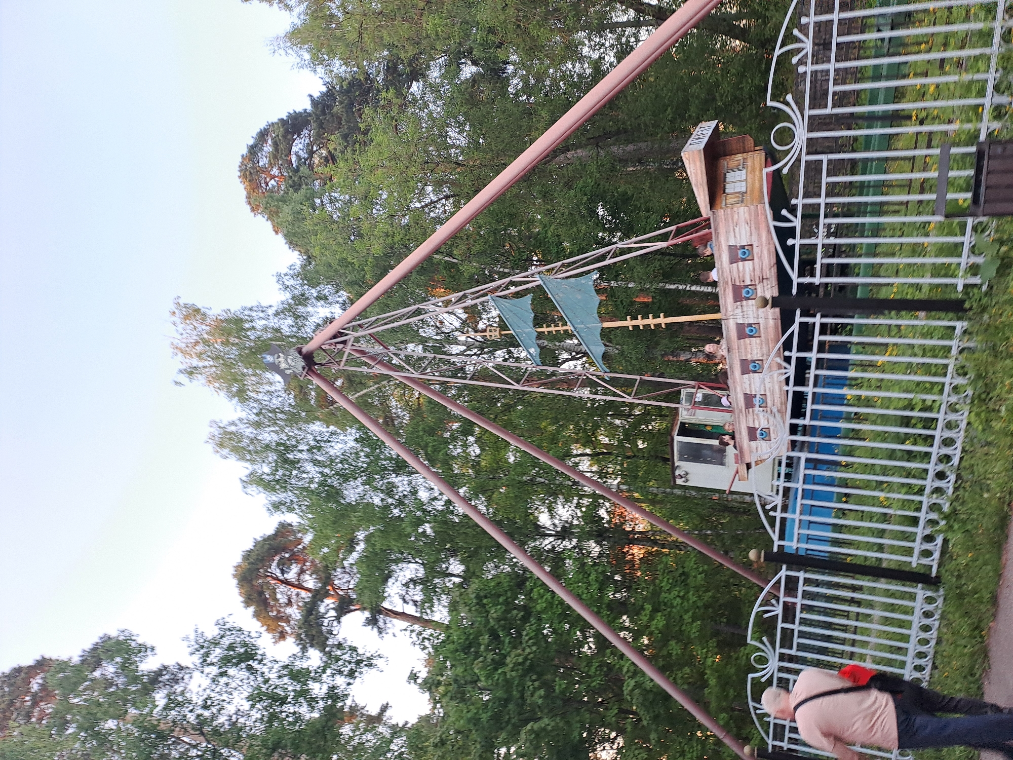 The image shows an amusement ride shaped like a ship, situated near a white metal fence. The ride has a wooden structure and what appear to be green sails. Tall trees are in the background. A person wearing a light-colored shirt and dark pants, with a red bag, is walking near the fence. The city is Noginsk, Russia.