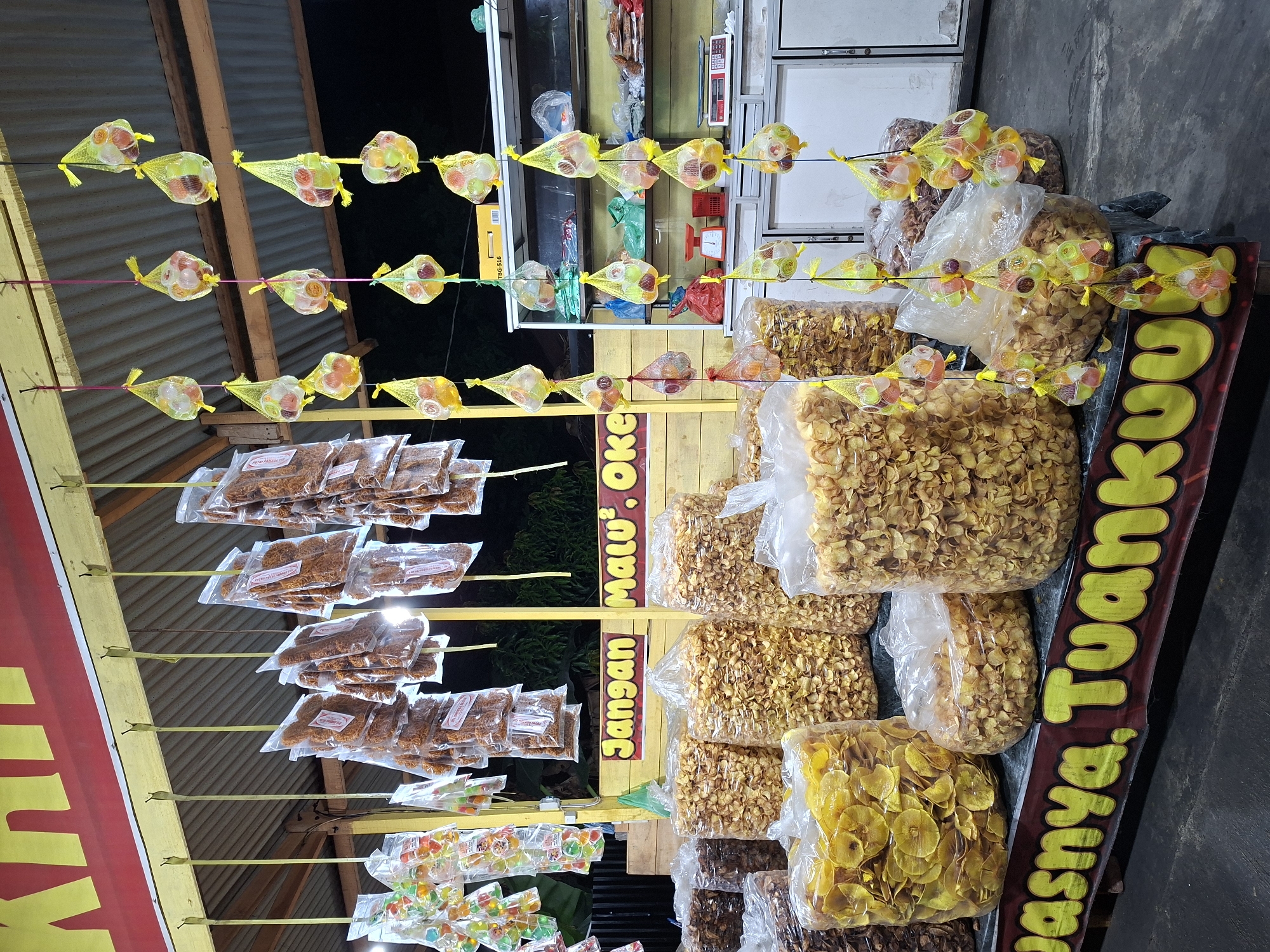 This image captures a nighttime food stall in Glee Gogo, Indonesia, displaying various snacks for sale. The scene is illuminated by artificial lighting, indicating operation after dark.
In the lower foreground, multiple large, clear plastic bags are filled with deep-fried chips; prominent among these are golden-yellow, thinly sliced circular chips, likely banana or cassava, alongside bags containing darker, irregularly shaped chips. A red banner with yellow, block-lettered text, partially visible at the bottom, reads "...asinya. Tuankuu."
Above these large bags, a yellow wooden beam supports numerous small, clear plastic bags containing reddish-brown, crispy savory snacks, possibly fried noodles or similar items, suspended by thin yellow ribbons. To the far left, additional small clear plastic bags containing assorted colorful jelly candies are also hung.
Centrally positioned, a yellow wooden structure frames a red banner with yellow, capitalized text stating "Jangan Malu'. OKE" (Don't be shy. OK).
To the right, a vertical arrangement of colorful jelly candies is displayed. Each candy, encased in a small clear cup, is individually held within a yellow mesh net and suspended by yellow and pink strings at various heights. Behind these hanging sweets, a white, multi-shelf cabinet is visible. The shelves hold various items, including plastic bags, a red mechanical weighing scale, and a modern digital scale. A yellow box marked "TBG-516" rests on top of the cabinet.
The background is predominantly dark, with hints of green foliage beyond the stall. The stall's roof is constructed from corrugated metal. No individuals are visible within the frame.