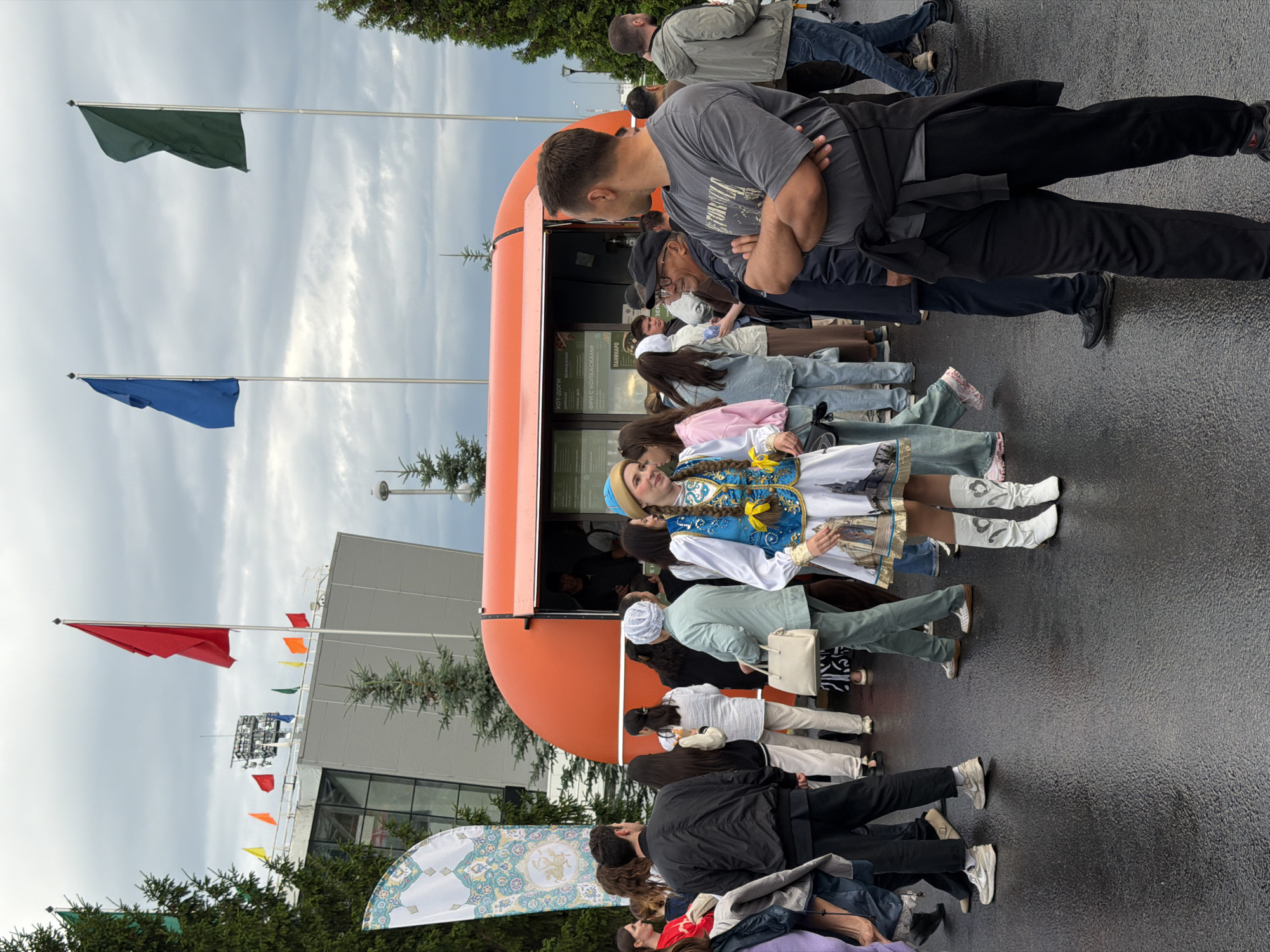 In a city square with a wet asphalt ground, a group of people are gathered around an orange food truck. The sky is overcast and cloudy, and there are tall green trees on the left and right sides of the image. Several flags are visible: red, yellow, orange, blue, and green flags on poles, as well as a large white banner with a decorative pattern on the left.
In the foreground, a young woman in a traditional blue and white outfit with gold embellishments and white boots is the most prominent figure. She is smiling and looking to her right, appearing to be interacting with someone. Behind her, a crowd of people are standing, some looking at the food truck menu, others conversing.
To the right of the food truck, a man in a gray t-shirt and black pants is standing with his arms crossed, facing away from the camera. Further to the right, another man in a gray hooded jacket and blue jeans is walking away from the food truck.
The food truck itself is orange and has a serving window where a person is visible. A menu is displayed on the side of the truck. The overall scene suggests an outdoor event or gathering.