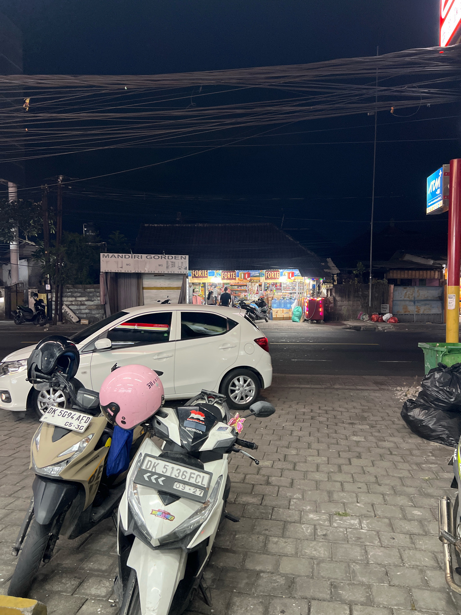 This is a nighttime street scene in Denpasar, Indonesia, featuring parked vehicles and a storefront. In the foreground, two scooters are parked side-by-side on a brick-paved sidewalk. The scooter on the left is beige and black, with a black helmet and a blue cloth hanging from its handlebars. Its license plate reads "DK 5G94 AFD 05.30". The scooter on the right is white and black, with a pink helmet and a sticker on its front that says "WONG JEMBER". Its license plate reads "DK 5136FCL 09.27". Behind the scooters, a white car is parked parallel to the street. The car has its headlights off.
Further down the street, a storefront is visible. It appears to be a small shop or convenience store, with bright interior lighting illuminating shelves stocked with various products. Signs with "FORTE" are visible on the storefront. People are seen inside and in front of the shop. Across the street, there are more buildings and some red plastic bags or trash piled up on the sidewalk next to a green garbage bin. The sky is dark, indicating it is nighttime, and a complex network of overhead wires is visible against the dark sky. The overall impression is a typical urban street scene in Indonesia at night, with the presence of shops, vehicles, and everyday urban elements.