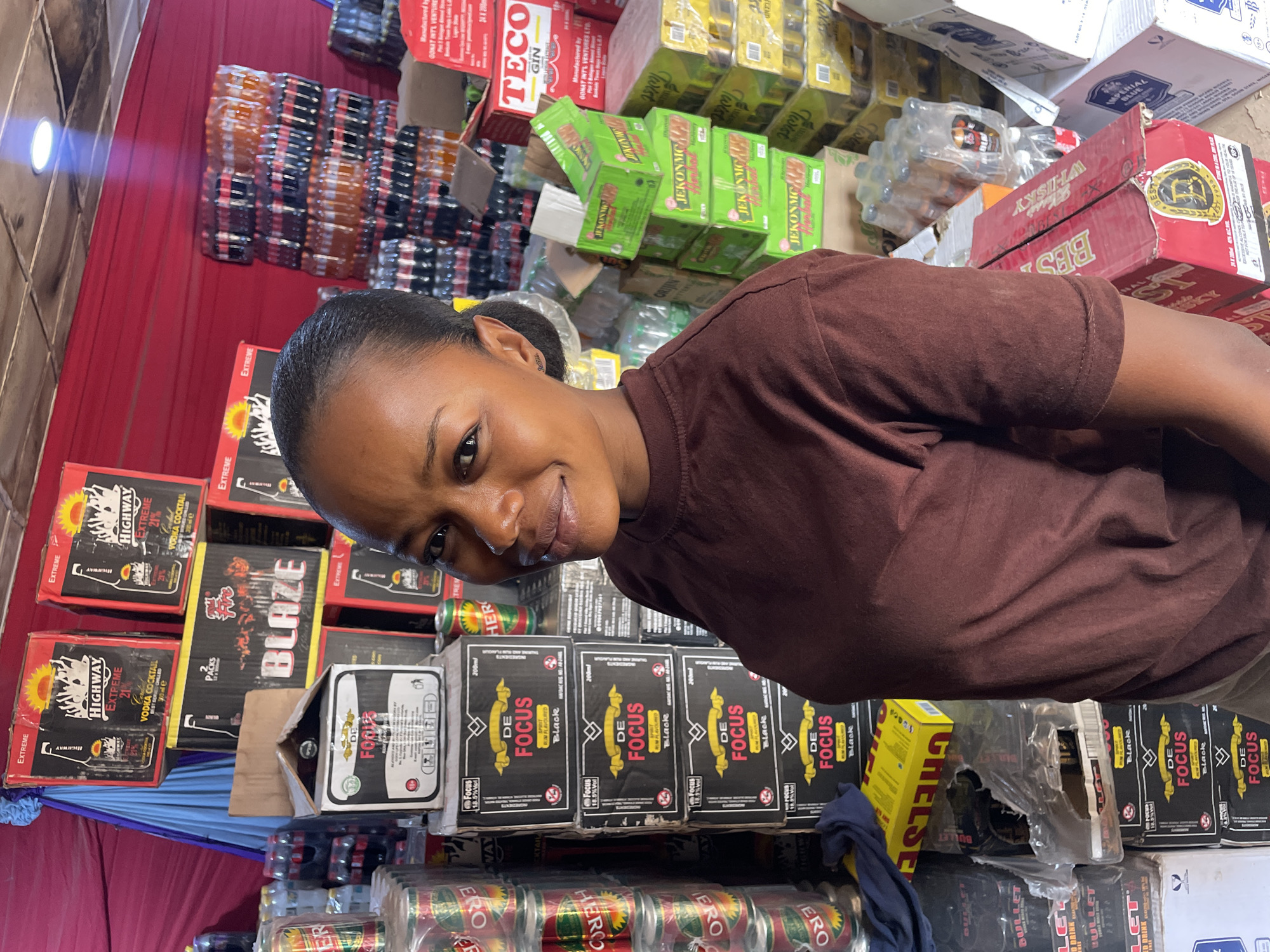 A young woman with dark hair pulled back and wearing a brown t-shirt is captured in a close-up, smiling slightly and looking directly at the camera. The setting is a cluttered indoor space, likely a beverage stockroom, wholesale store, or a very well-stocked shop in Jos, Nigeria.
The background is densely packed with a large assortment of stacked boxes, cartons, and cans of various beverages. These items form a wall behind and around the woman, suggesting a commercial environment focused on drink sales or distribution. A bright light source is visible on the far left of the image.
Visible text on the product packaging includes brand names such as "DE FOCUS Black" (an 18.5% Vol spirit drink), "HIGHWAY EXTREME" (a 21% Vodka Cocktail), "BLAZE," "TECO GIN," "JEKONMC," "BESTO WHISKY," "IMPERIAL BLUE," "CHELSE," "BULLET" (energy drink), "HERO," "ERO," and "LET ODRINK." Some packaging specifies "200ml" content. One label mentions "Manufactured by GOKAT INT'L VENTURES LTD." The activity appears to be a casual photograph, possibly a selfie, within this busy retail or storage environment.
