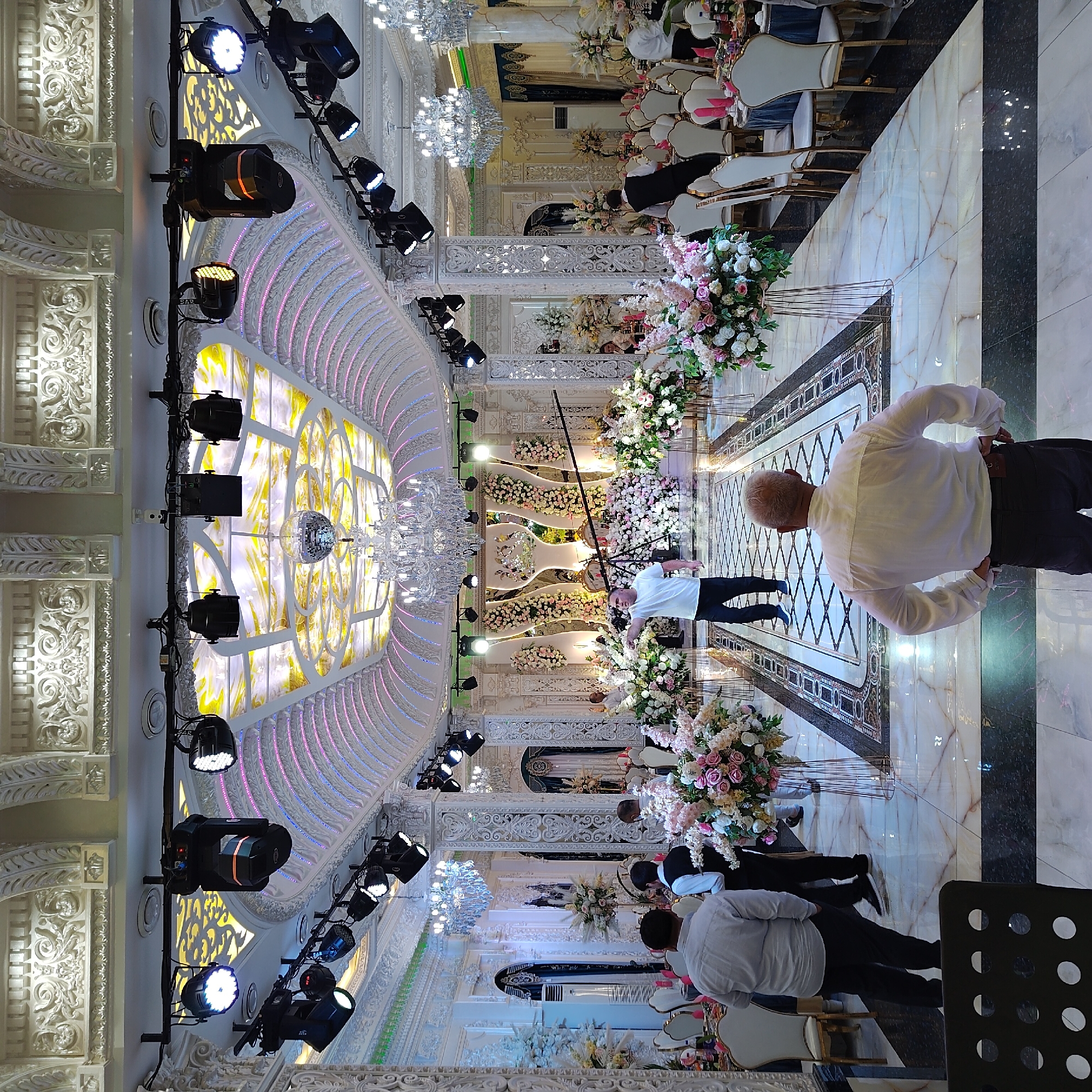 The image depicts the interior of a richly decorated banquet hall, likely in Khujand, Tajikistan, set up for a formal event. The hall features ornate white plasterwork on the ceilings and walls, elaborate chandeliers, and a central mirrored disco ball. Spotlights are mounted on overhead trusses, suggesting a performance or entertainment is planned. Floral arrangements of pink and white flowers are prominent, placed on tall metallic stands along a decorative tiled aisle. Tables are set with white chairs, some draped in blue tablecloths, indicating dining arrangements. Several individuals are visible throughout the hall; one man in a white t-shirt and dark pants is standing in the aisle facing the stage area, gesturing with his left hand. Another man in a white shirt and dark pants stands with his back to the camera, hands on his hips, facing the aisle. Other figures, some in formal attire, are seen in the background near the tables. The flooring is a polished marble with intricate geometric patterns. The overall impression is one of opulence and preparation for a celebratory gathering.