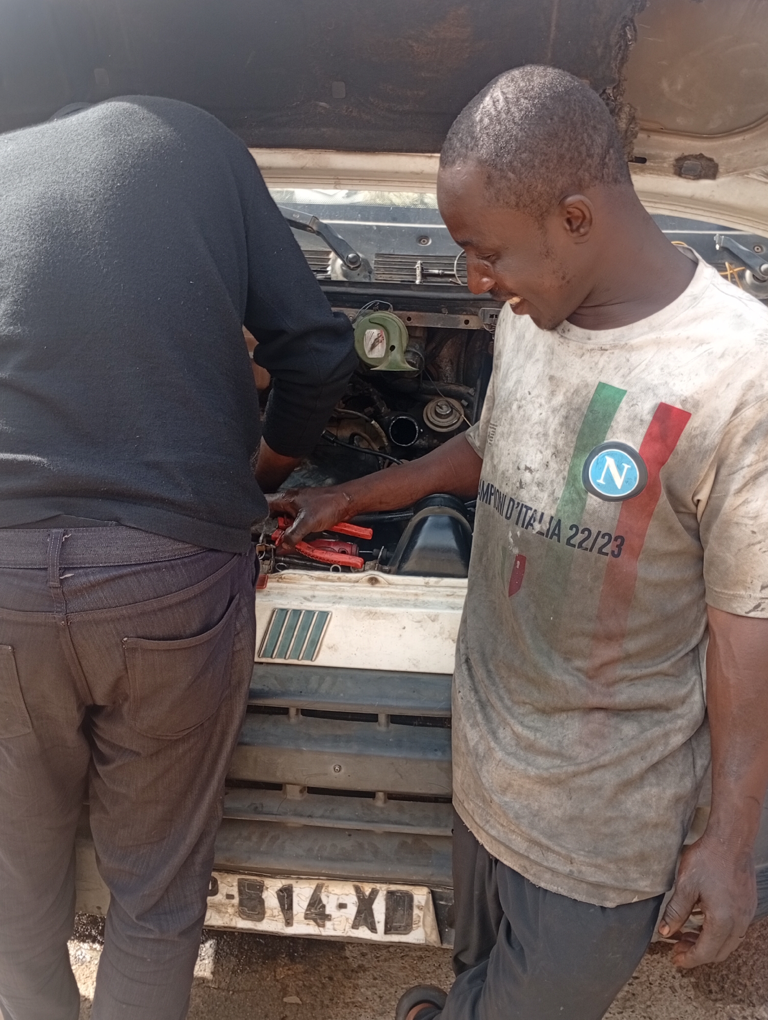 Two individuals are seen working on the engine of a vehicle, likely in an outdoor setting in Rafin Par, Nigeria. The person on the right, wearing a dirt-stained t-shirt with Italian flag colors and a Napoli logo, is actively engaged with the engine, holding a pair of red pliers. Their expression suggests focus and concentration. The other individual, to the left and mostly obscured from the chest down, wears a dark long-sleeved shirt and dark pants, also appearing to be working on the engine.
The scene suggests a mechanical repair or maintenance activity. The vehicles engine compartment is open, revealing various engine components. The ground appears to be dirt or gravel. The lighting suggests it is daytime, with natural sunlight illuminating the scene. The overall atmosphere is one of practical work.
Visible text includes "CHAMPION D'ITALIA 22/23" on the t-shirt of the person on the right, along with the Napoli crest. There is also a license plate on the front of the vehicle with the characters "P-B14-XD". The environment appears to be a functional area for vehicle repair, possibly a roadside workshop or a personal garage.
