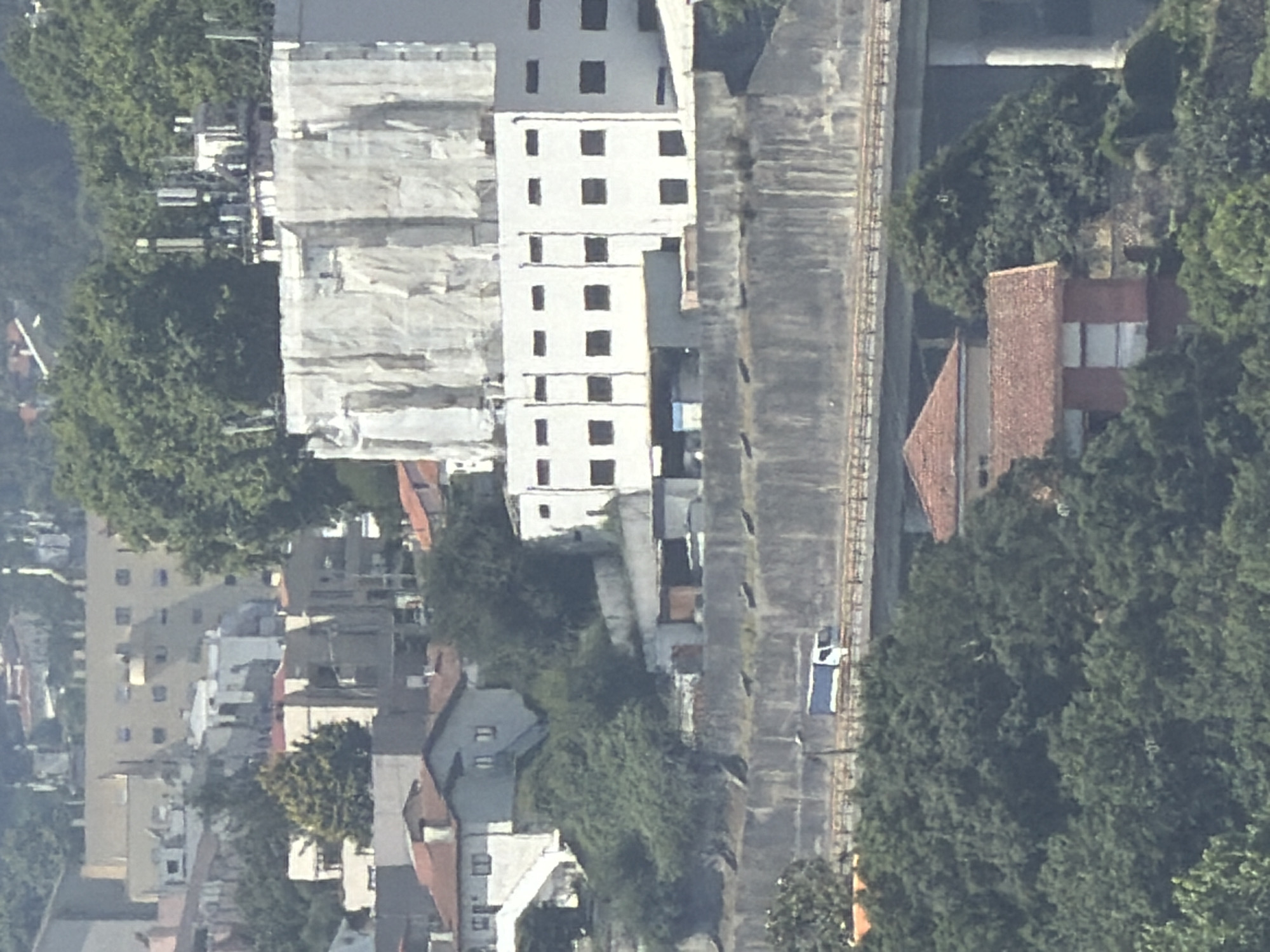 This aerial view captures a dense urban and natural landscape in Gondomar, Portugal, likely during daylight with clear weather. The scene is dominated by a cluster of buildings, some appearing under construction or renovation due to scaffolding and white tarps covering large sections. A tall, multi-story white building with numerous dark, rectangular windows stands prominently.
Adjacent to the buildings is a long, grey concrete structure that appears to be a raised walkway or elevated platform, possibly a viaduct or embankment. A thin, dark line running along its length suggests tracks or a similar linear feature. A small, white vehicle with a blue stripe is visible on this elevated structure.
Further into the scene, traditional Portuguese architecture is evident with buildings featuring red-tiled roofs. Dense green foliage, indicative of trees and vegetation, is interspersed throughout the urban area, suggesting a well-established park or residential gardens. The overall impression is one of a developed area with a mix of modern and older structures, partially integrated with natural elements. No people are clearly discernible, and no text is visible in the image.