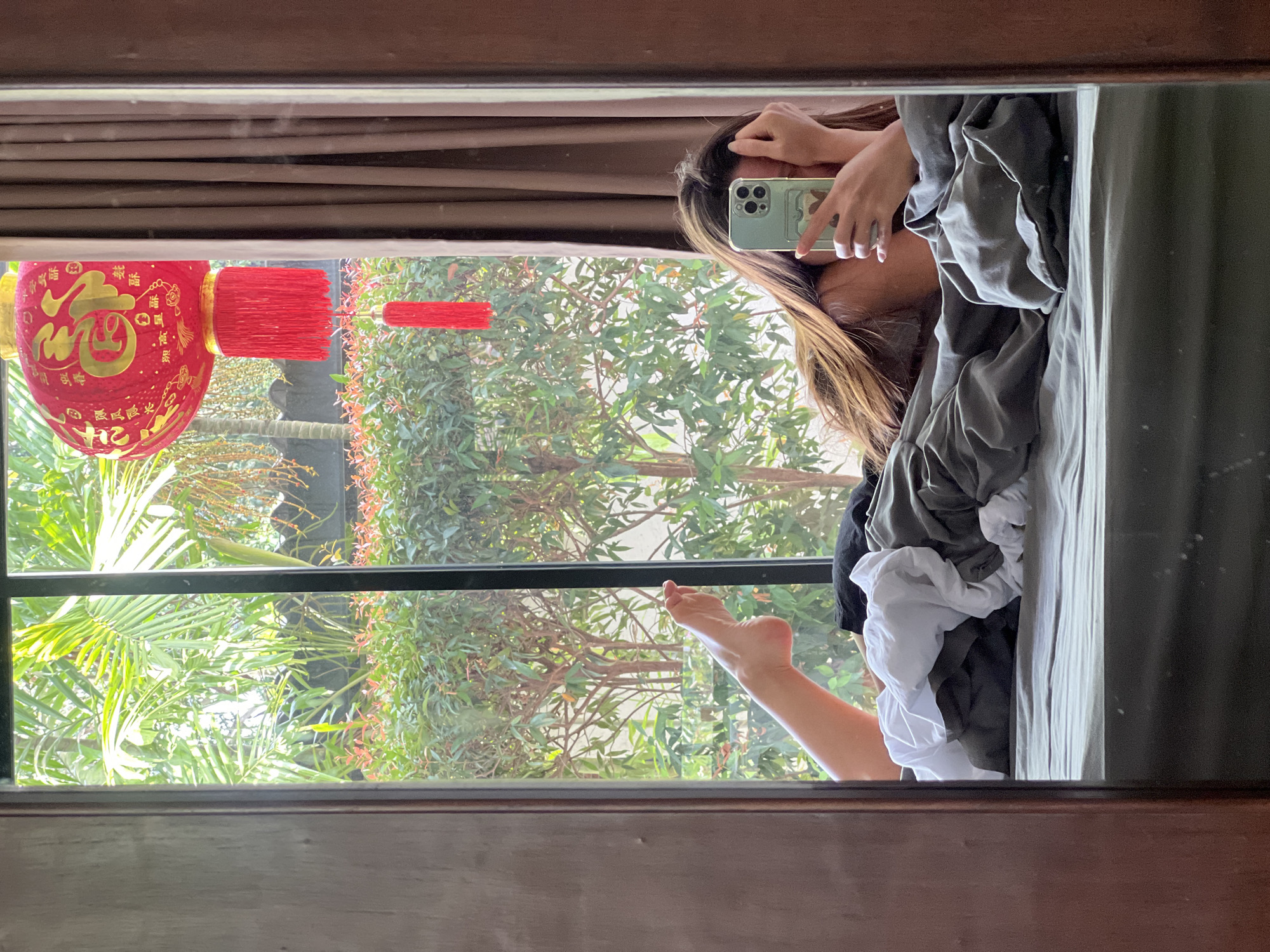A mirror selfie of a person lying on a bed with a phone in their hand. The reflection shows a view of lush greenery with a red Chinese lantern hanging outside the window. The person's foot is extended toward the window, and they are partially covered by bedclothes. The location is Unknown, Unknown.
