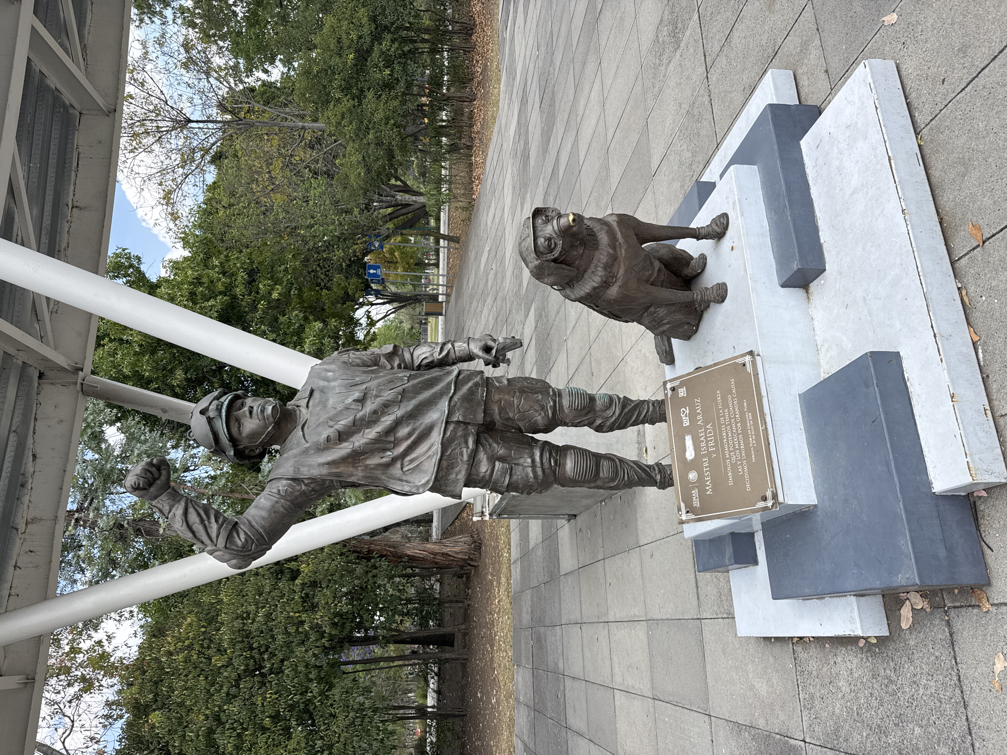 This photograph shows a bronze sculpture of a man and a dog in a park setting. The man is depicted wearing a helmet, goggles, and riding gear. He is standing with his fist raised in the air. Next to him, on a raised platform, sits a dog wearing goggles. The sculpture is mounted on a stepped concrete base with a plaque attached. The plaque identifies the sculpture as "MAESTRE ISRAEL ARAUZ Y FRIDA" and states that they are memorable symbols of the strength that Mexicans can have when they unite for great causes. The plaque notes that the sculpture is from Zaragoza Puebla and was dedicated in July 2018.
The sculpture is located on a tiled walkway in the park. Behind the sculpture, there are trees and vegetation. To the left, there is a white architectural structure, possibly a covered walkway. In the background, other trees and foliage can be seen. The statue is dedicated to Israel Arauz and a rescue dog named Frida, and is in Puebla, Mexico.