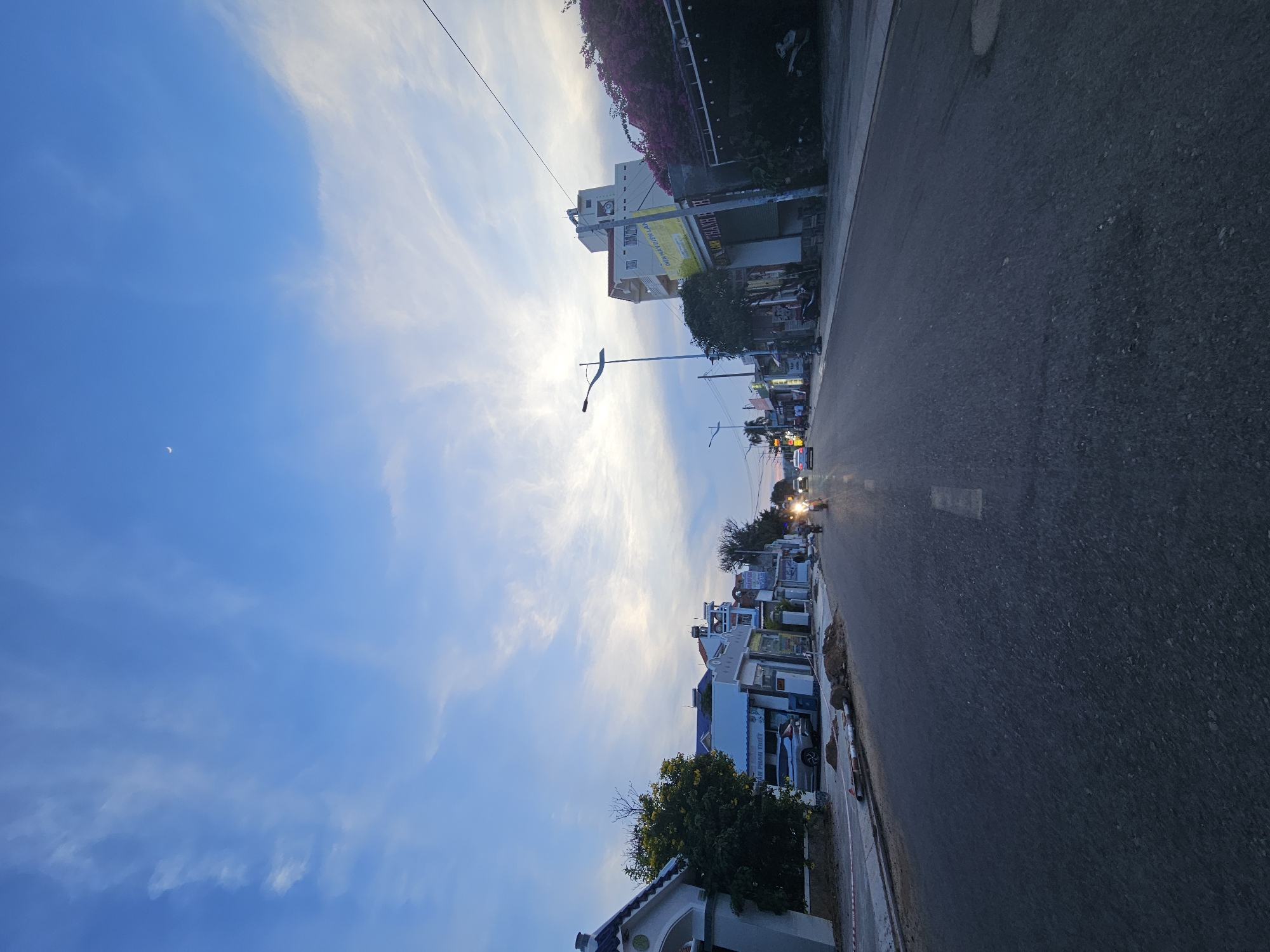 The image depicts a street scene in Phu Thuy Ward, Vietnam, during what appears to be late afternoon or early evening, given the soft light and the presence of a crescent moon in the sky. The sky is a vibrant blue, with wispy white clouds stretching across it. The perspective is from a low angle, looking down a paved road lined with buildings.
The buildings on the left side of the road vary in height and style, with some appearing to be multi-story residences or small businesses. One building prominently displays a yellow sign with text in Vietnamese, which is partially legible. Other signs and advertisements are visible on the building facades, suggesting a commercial or residential area. There are trees and some flowering bushes, one with vibrant pink flowers, adding touches of nature to the urban landscape.
Several vehicles are present on the road and parked along the side. A motorcycle with its headlights on is the most prominent vehicle, approaching the viewer. Other cars and motorcycles are also visible, indicating typical street activity. The road itself shows signs of wear, with some patches and imperfections.
The overall atmosphere is tranquil, with a sense of everyday life unfolding. The lighting suggests a transition from day to night. The location cues, such as the architecture and the Vietnamese text, firmly place the scene in Vietnam. No people are distinctly identifiable in the image. The image is captured with the camera oriented vertically.