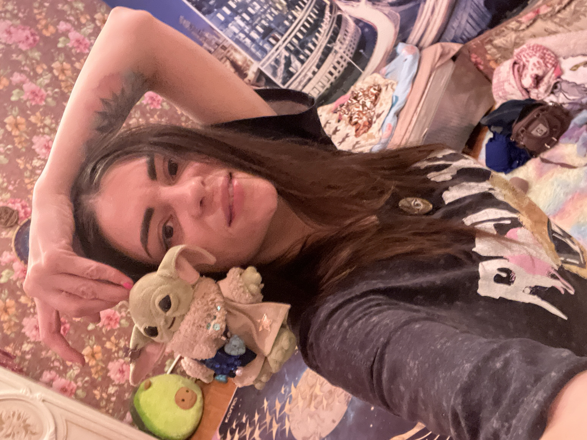 The image depicts a close-up selfie of a young woman holding a plush toy resembling Grogu from Star Wars. She has long, dark hair, dark eyebrows, and is wearing a dark grey t-shirt with a graphic print. Her right arm is raised, and her hand is gently touching the top of the toy's head. To her left, there is a plush toy shaped like an avocado. The background features a floral patterned wallpaper and what appears to be a bed with various blankets and textiles. There are no discernible indicators of the specific location within the city of Krasnoe-na-Volge, Russia. The scene appears to be indoors, likely in a bedroom.