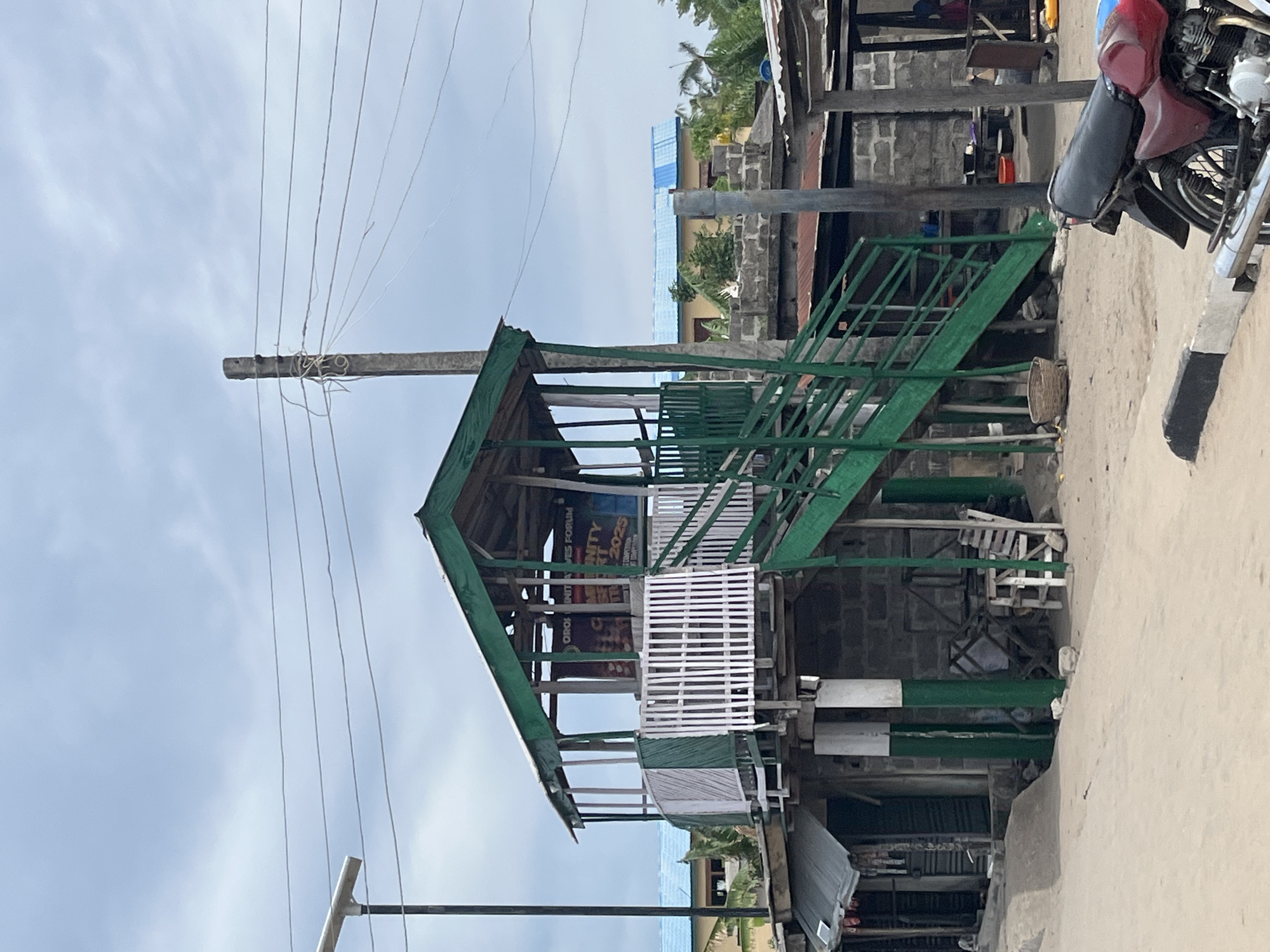 The image shows a two-story wooden structure in Badagry, Nigeria. The structure is painted green and white. It features stairs, railings, and a roof. A poster is visible inside the structure. Power lines are strung above the building from a pole. A red motorcycle is parked nearby on a sandy ground. Adjacent to the structure, there are other buildings made of concrete blocks and corrugated iron. The sky is overcast.