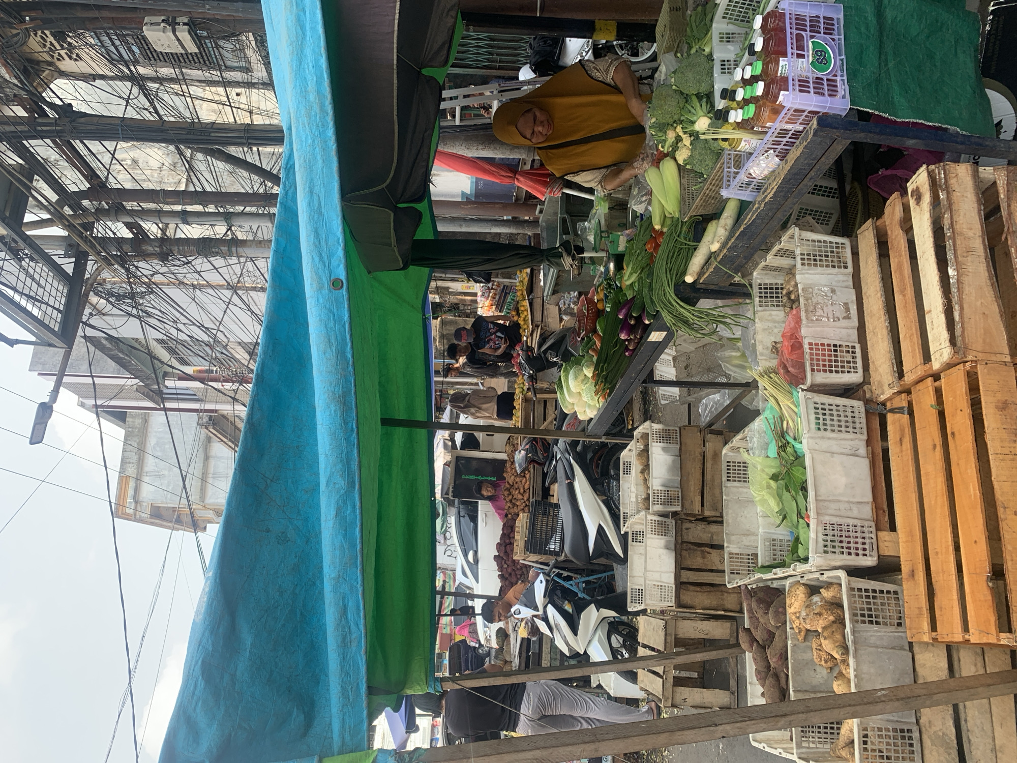 This image depicts a bustling street market scene in Palembang, Indonesia, likely in a neighborhood like 10 Ilir, known for its vibrant markets. A vendor, wearing a yellow hijab and a light brown patterned top, is busy arranging produce at a stall. Various fresh vegetables, including broccoli, cucumbers, long beans, and leafy greens, are displayed in white plastic crates. There are also bottles of drinks in a purple crate on a metal rack. In the background, several motorcycles are parked, and other vendors and customers are visible, suggesting a busy commercial area. The market is covered by blue and green tarpaulins, providing shade from the sun. The urban environment is characterized by buildings and an abundance of overhead electrical wires, typical of densely populated city areas in Indonesia. The overall atmosphere is one of everyday commerce and community interaction.