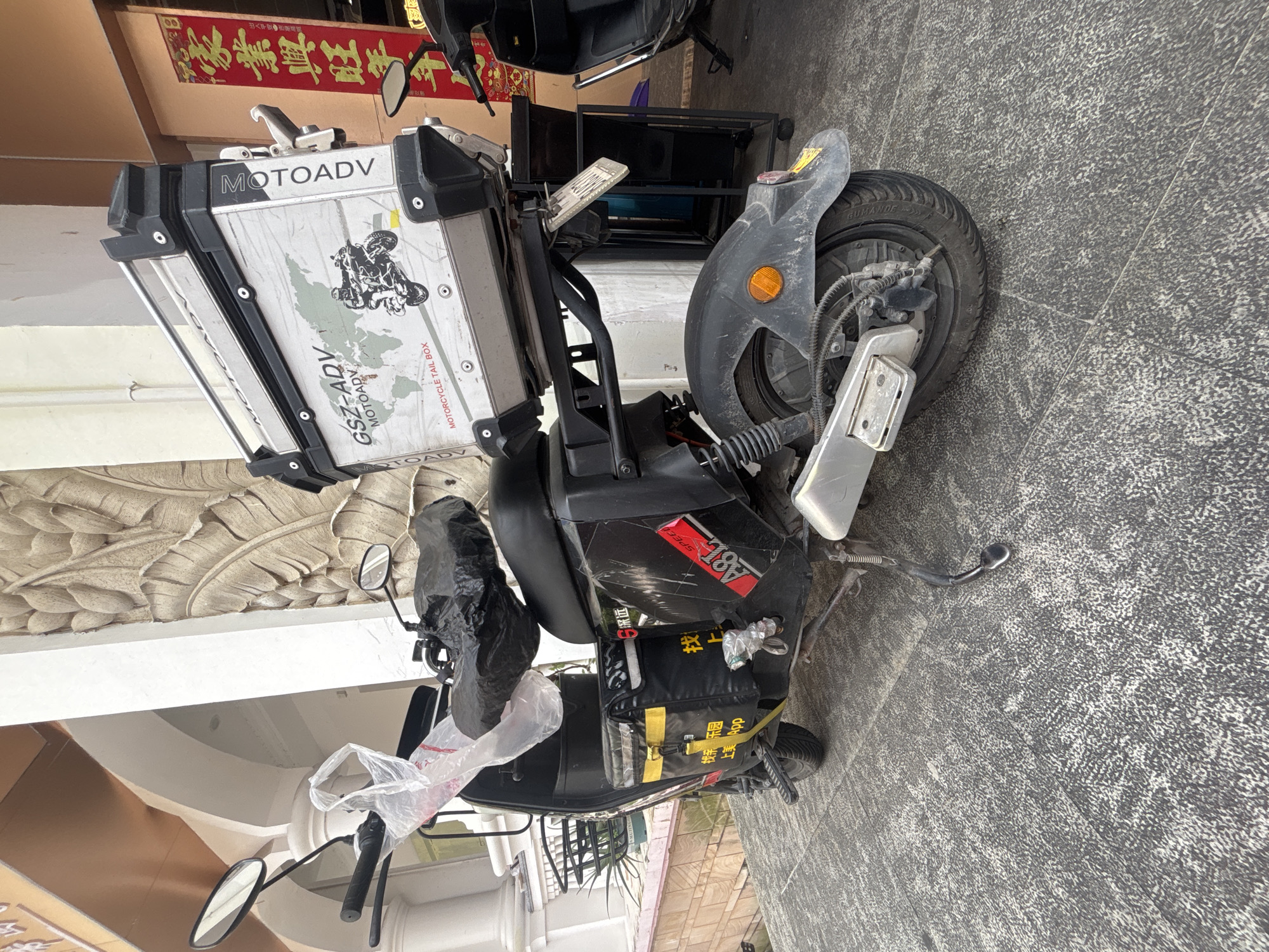 An electric scooter is parked on a tiled pathway in front of a building in Nanhai District, China. The scooter is black and equipped with a large silver storage box labeled "MOTOADV GSZ-ADV" on its rear. The storage box features a graphic of a world map and a motorcycle. On the side of the scooter, there is a black delivery bag with yellow trim and Chinese characters. A plastic bag and a black cover are placed on the handlebars and seat, respectively. The building behind the scooter has an arched entrance and decorative relief carvings on its wall. A red banner with Chinese calligraphy is visible to the right. The ground is paved with textured gray tiles. Another scooter is partially visible in the background to the right.