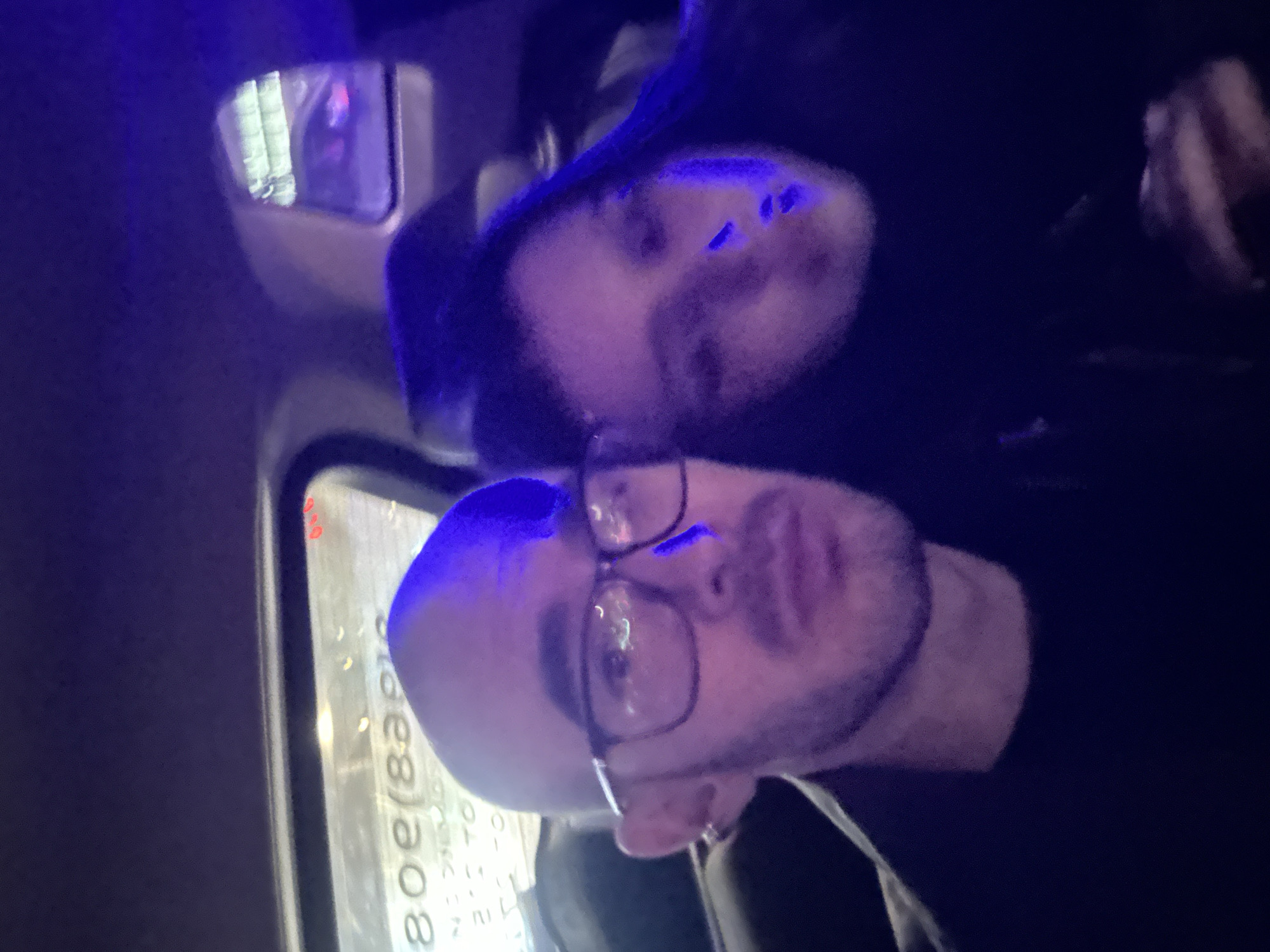 Two people are in a vehicle. One person is wearing glasses, and the other is behind. A side mirror is visible with reflections. A window is also visible with text reading "080(839". The overall lighting has a blue tint. The location is likely Moscow, Russia, as indicated by the phone number prefix.