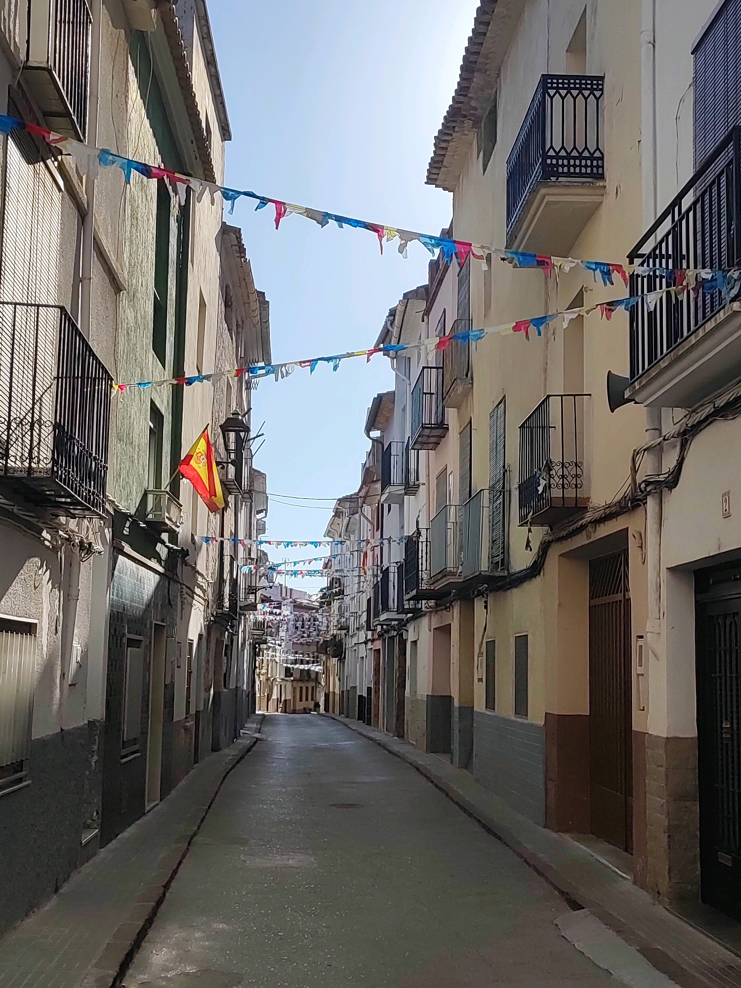 This is a street-level, eye-level, medium shot of a narrow street in Sueras, Spain. The street is lined with buildings on both sides, with balconies on some of the upper floors. Colorful flags are strung across the street between the buildings. A Spanish flag is visible on the left side of the street. The street is paved and appears to be wet, reflecting the light. The sky is clear and blue, with bright sunlight casting shadows. The scene appears to be during daytime.