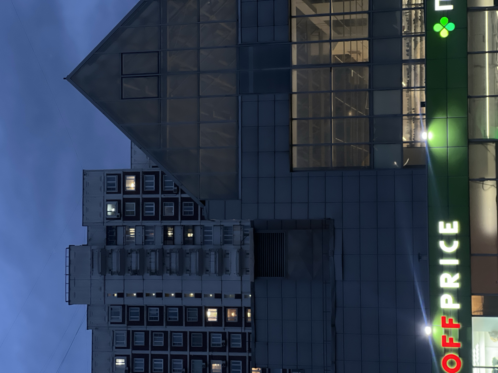 The image shows a building complex in Moscow, Russia, at twilight. A high-rise apartment building with numerous windows, some lit, is adjacent to a commercial building with a triangular architectural element and large windows. The commercial building has a prominent "OFF PRICE" sign. The sky is a dusky blue.