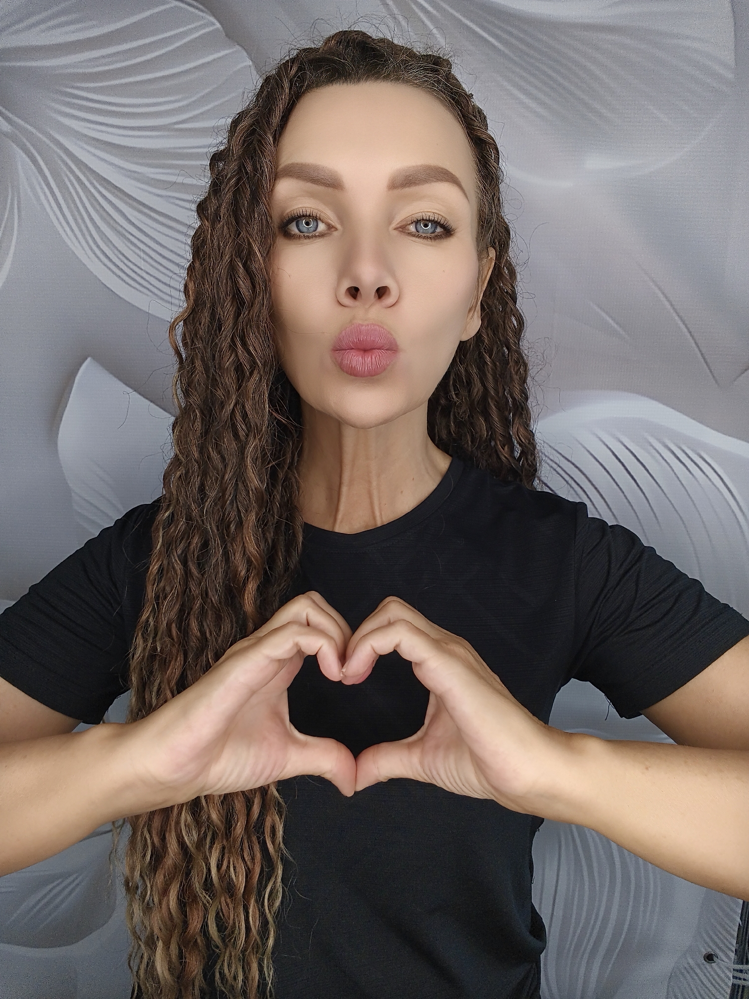 A young woman with long, curly brown hair and bright blue eyes is posing for a photo. She is wearing a black t-shirt and making a heart shape with her hands. Her lips are pursed, as if she is blowing a kiss. The background is a light gray wall with a floral pattern. The woman is positioned in the center of the frame, with her head slightly tilted. Her expression is playful and inviting. The lighting is soft and even, highlighting her features. The image appears to be a selfie taken indoors.