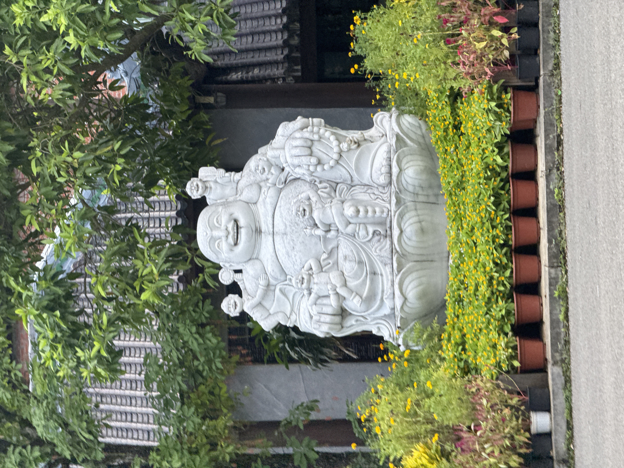 In Tây Hoa Lư Ward, Vietnam, a stone statue of a laughing Buddha, also known as Budai, is prominently displayed outdoors. The statue depicts a plump, jovial figure seated on a lotus pedestal, surrounded by several cherubic children. The Buddha statue is the central element of the scene, positioned in front of a building with a traditional tiled roof and lush green trees. The foreground is adorned with a vibrant arrangement of potted flowers, including rows of small yellow blooms and some taller plants with pinkish-red foliage. The ground in front of the potted plants is a paved surface, likely a walkway or street. The overall setting appears to be an outdoor space, possibly a garden or a courtyard area of a temple or cultural site.
