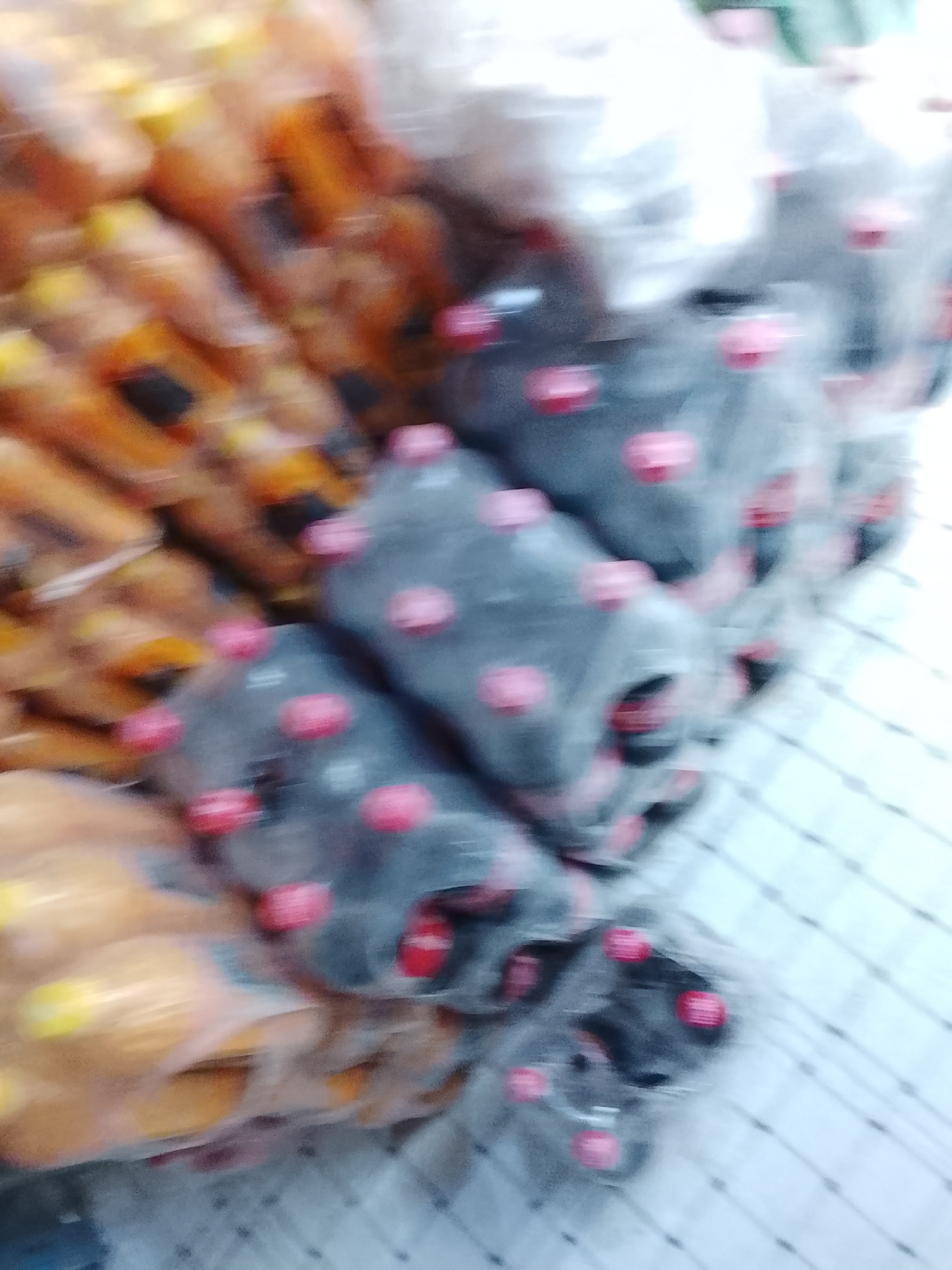 The image, taken in Gulgasht Colony, Pakistan, shows stacks of bottled beverages on a checkered surface. Bottles with orange liquid and yellow caps are stacked to the left, while dark liquid in bottles with pink caps are piled to the right. The image is blurry, obscuring specific details.