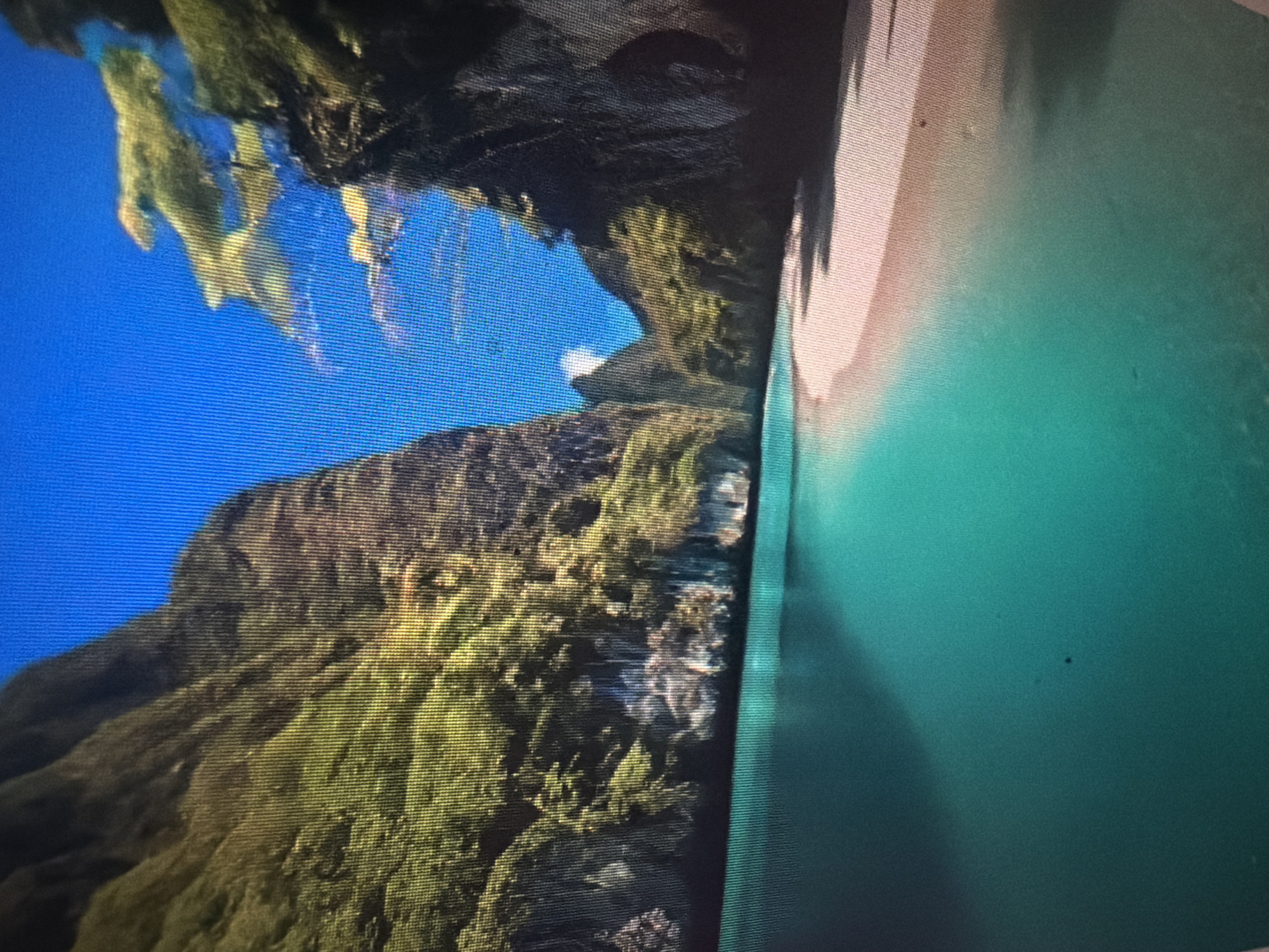 This is a digitally rendered image depicting a tropical beach scene with dramatic limestone cliffs flanking a body of turquoise water and a sandy shoreline. The sky is a vibrant, cloudless blue. The cliffs are covered in lush green vegetation, suggesting a tropical environment. The water is clear and calm, with subtle reflections of the cliffs and sky. The beach appears to be composed of fine, light-colored sand, with some shadows cast by the cliffs. The image evokes a sense of secluded paradise. There is no indication of any specific location within the city of Baan Mae Nam, Thailand, or any people present.