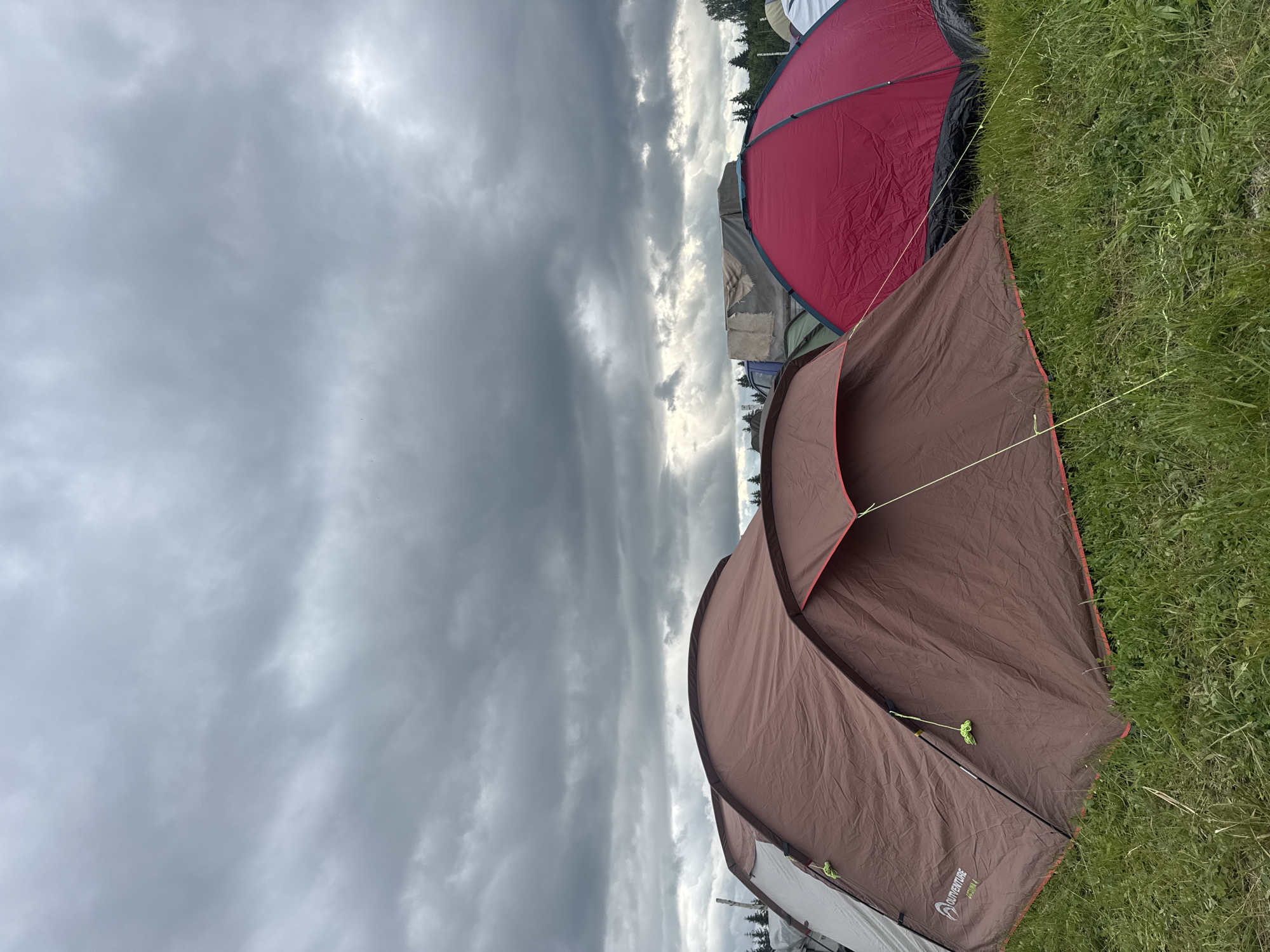 The image presents an outdoor camping scene in Bolshaya Kivara, Russia, under a heavily overcast sky. The upper two-thirds of the frame are dominated by dark, dense, charcoal-grey clouds, with a few slightly brighter patches indicating diffused light, particularly towards the right. A line of dark green coniferous trees is visible along the distant horizon, partially obscured by various tents and makeshift structures.
In the midground and background, numerous other tents of varying colors (green, white, dark blue) are set up, some partially covered by tarps, suggesting a crowded encampment. A blue vehicle and a large structure covered by a torn grey tarp are visible behind the tents in the center-left.
The foreground features two prominent tents pitched on a field of green grass with some lighter, drier patches. On the left, a large, dome-shaped brown (maroon/sepia) 'OUTVENTURE ZEPHYR 4' tent stands, displaying light grey panels on its left side and highlighted with bright yellow-green guy lines and red trim along its base and openings. To its immediate right, partially obscured, is a vivid red dome tent with a black base and green structural elements.
No individuals are visible, nor are any specific actions or interactions depicted. The environment suggests a temporary settlement or festival ground.