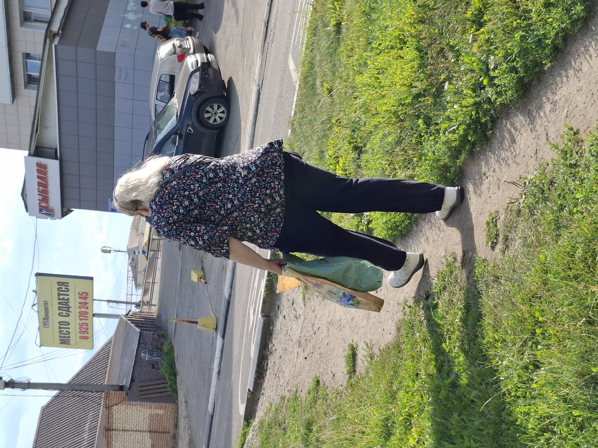 In a sunlit outdoor scene in Unknown, Unknown, a woman with blonde hair, wearing a dark floral print blouse and dark pants, walks away from the camera on a dirt path lined with green grass. She carries two large shopping bags, one green and one orange, in her left hand. To her right, a paved road runs parallel to the path, with several parked cars visible. Across the road, a building with a sign that reads "РЫБОЛОВ" (Rybolov, meaning Fisherman) is visible, as is a large yellow billboard with Russian text and a phone number. The sky is partly cloudy.