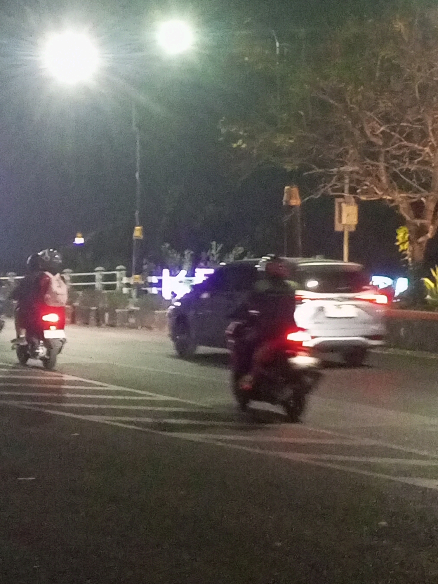 The image depicts a nighttime scene on a street in Kediri City, Indonesia. Two motorcycles and a car are visible. The foremost object is a motorcycle with a rider wearing a helmet, moving away from the camera. Its taillight is illuminated. To the left, another motorcycle with a rider is also visible. Further in the background, a car is moving away from the camera, with its taillights illuminated. The environment includes streetlights, trees, and a roadside barrier. The street is paved and appears to have lane markings. The overall impression is that of traffic on a city street at night.