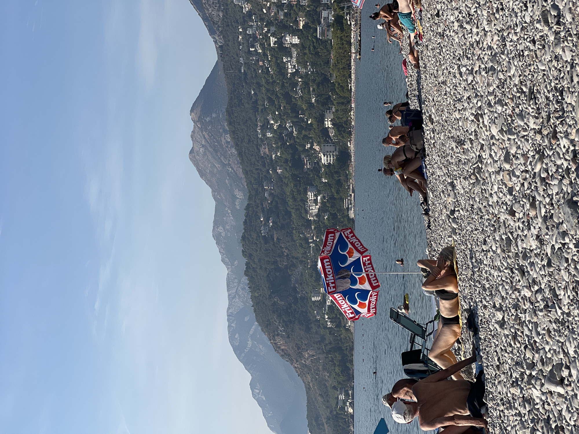 An outdoor, eye-level shot shows a crowded pebble beach with the Adriatic Sea in the background and mountains overlooking the city of Bar, Montenegro. The water is calm with a few swimmers visible. A colorful umbrella is in the foreground. People are sunbathing on towels and lounge chairs, some are seated in groups. The sky is clear and blue with some distant clouds.