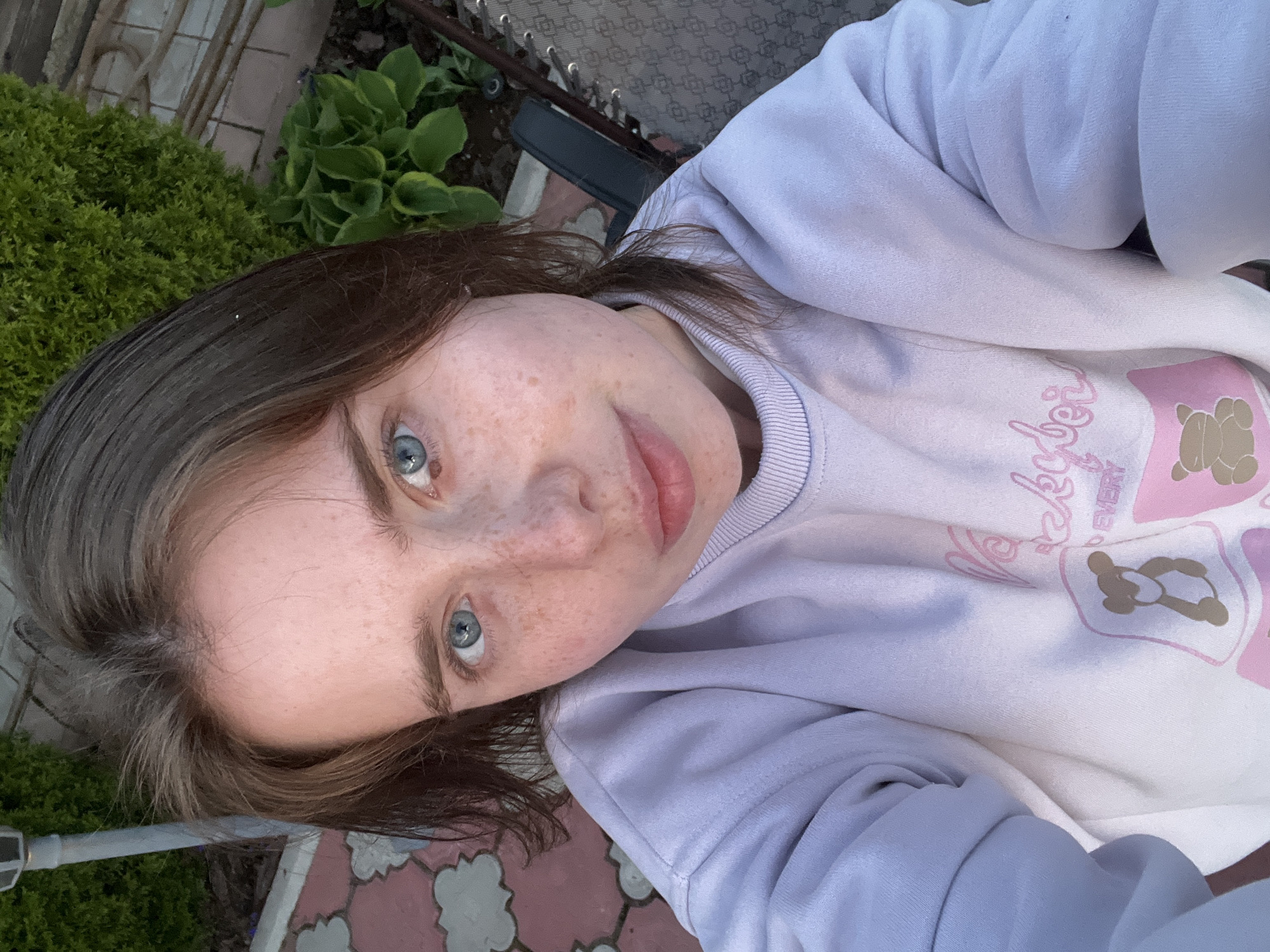 A close-up, slightly angled, selfie-style shot of a woman with fair skin, freckles, and light blue eyes. She has medium-length brown hair. She is wearing a white and light purple sweatshirt with a graphic design featuring a teddy bear and text.
The background includes greenery and paving stones. A portion of a chair is also visible. The location is inferred to be Chaikovsky, Russia, based on the prompt, though nothing in the image confirms this.