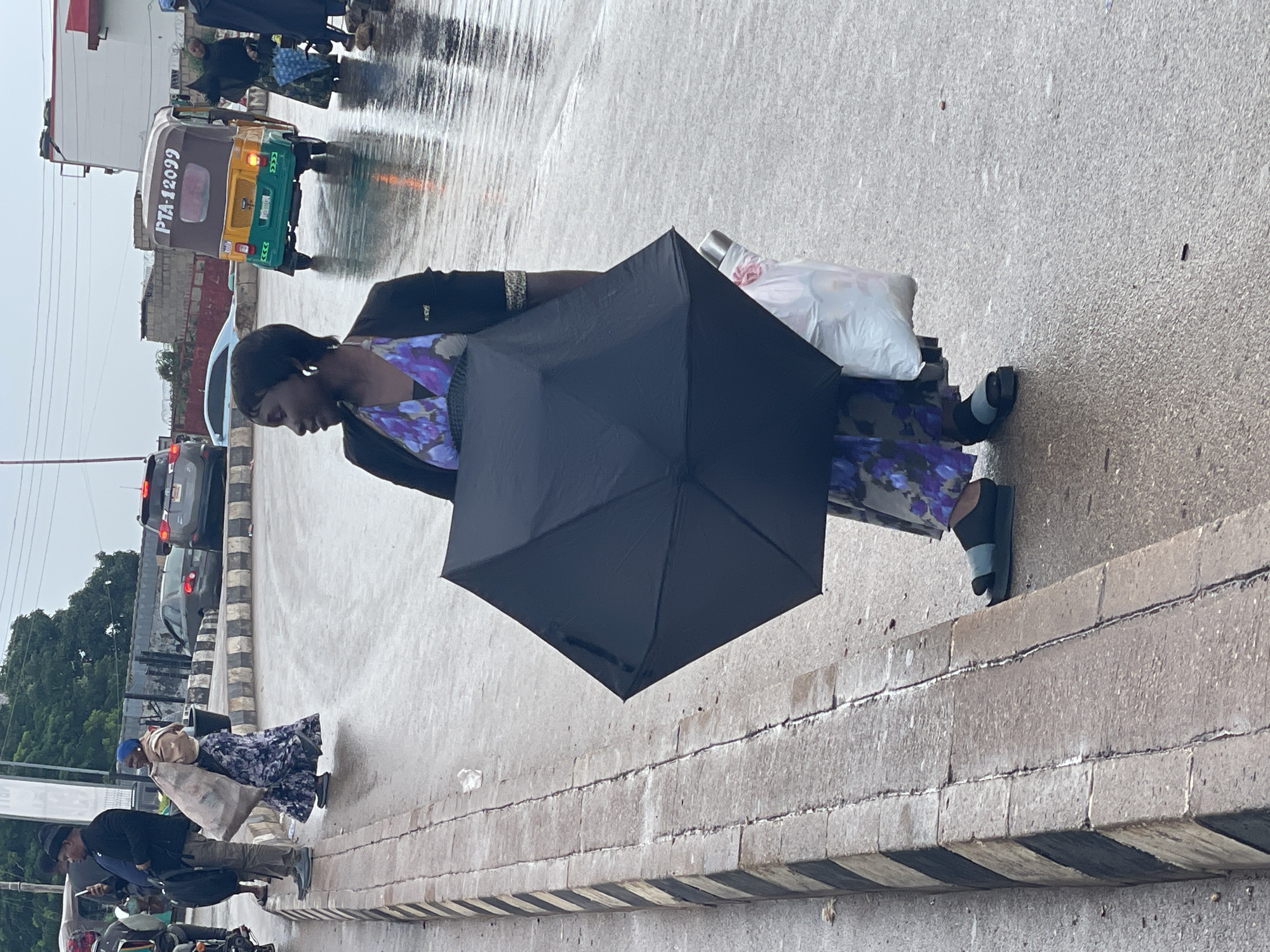A medium shot captures a woman holding a black umbrella while standing on a wet street in Jos, Nigeria. She is wearing a dark jacket over a purple and blue floral dress and dark sandals. The street is wet, indicating rain, with reflections visible on the surface. To her left, a striped concrete curb runs along the edge of the sidewalk. In the background, a yellow and green auto-rickshaw with the license plate "PTA-12099" is driving away. Other vehicles and pedestrians are visible in the distance, with trees and buildings forming the backdrop under an overcast sky. The scene appears to be a typical street view in the city.