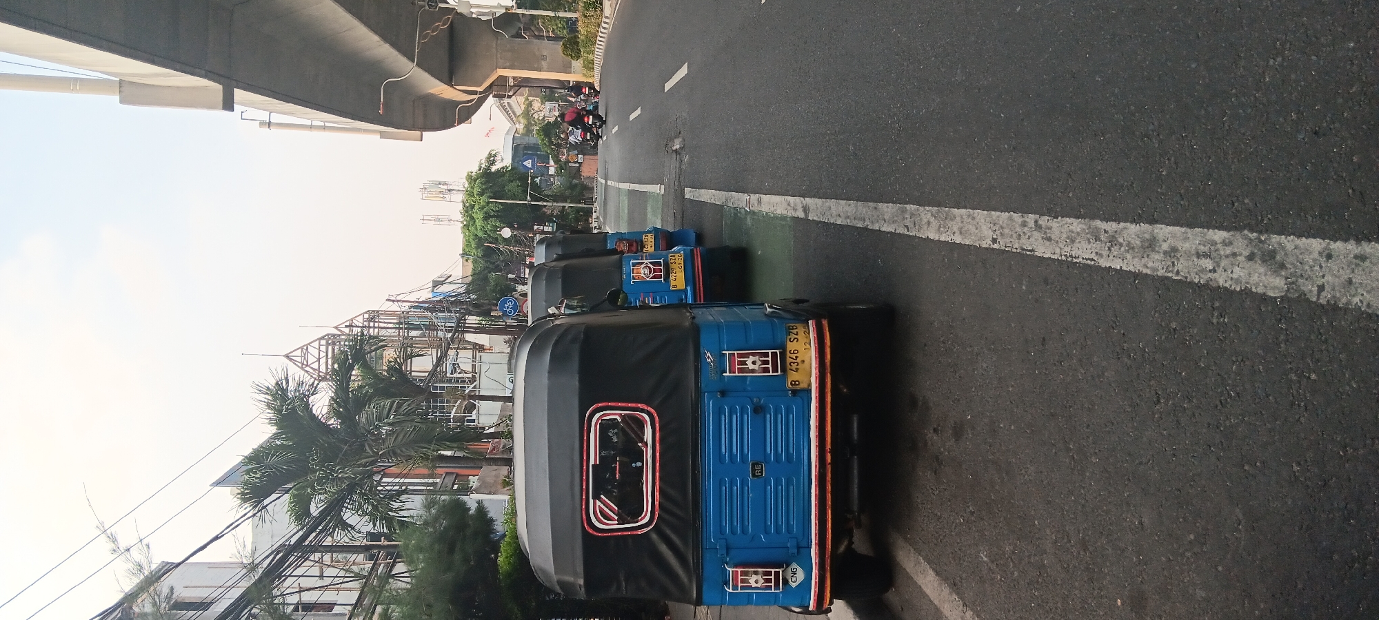 The image captures a street scene in South Jakarta, Indonesia, featuring a row of three blue autorickshaws (known locally as "bajaj") parked along the side of a road. The bajaj are predominantly blue with black canopies. The closest bajaj has a yellow license plate reading "B 4346 SZB" and "12-25" below it. The second and third bajaj are partially visible behind the first, with their license plates also visible but less clear.
The road itself is asphalt, with a white lane marking running parallel to the parked bajaj. To the right of the road, an elevated concrete structure, likely part of an overpass or highway, dominates the upper right portion of the frame. Beyond the overpass, there are buildings and trees. On the left side of the road, there are more buildings, palm trees, and a complex network of electrical wires and poles, characteristic of urban infrastructure. The sky is overcast with a light haze.
In the distance on the road, several motorcycles are visible, indicating some traffic. One motorcycle carries a passenger, and another appears to have two riders. The overall impression is of a typical urban street environment in Indonesia during daylight.