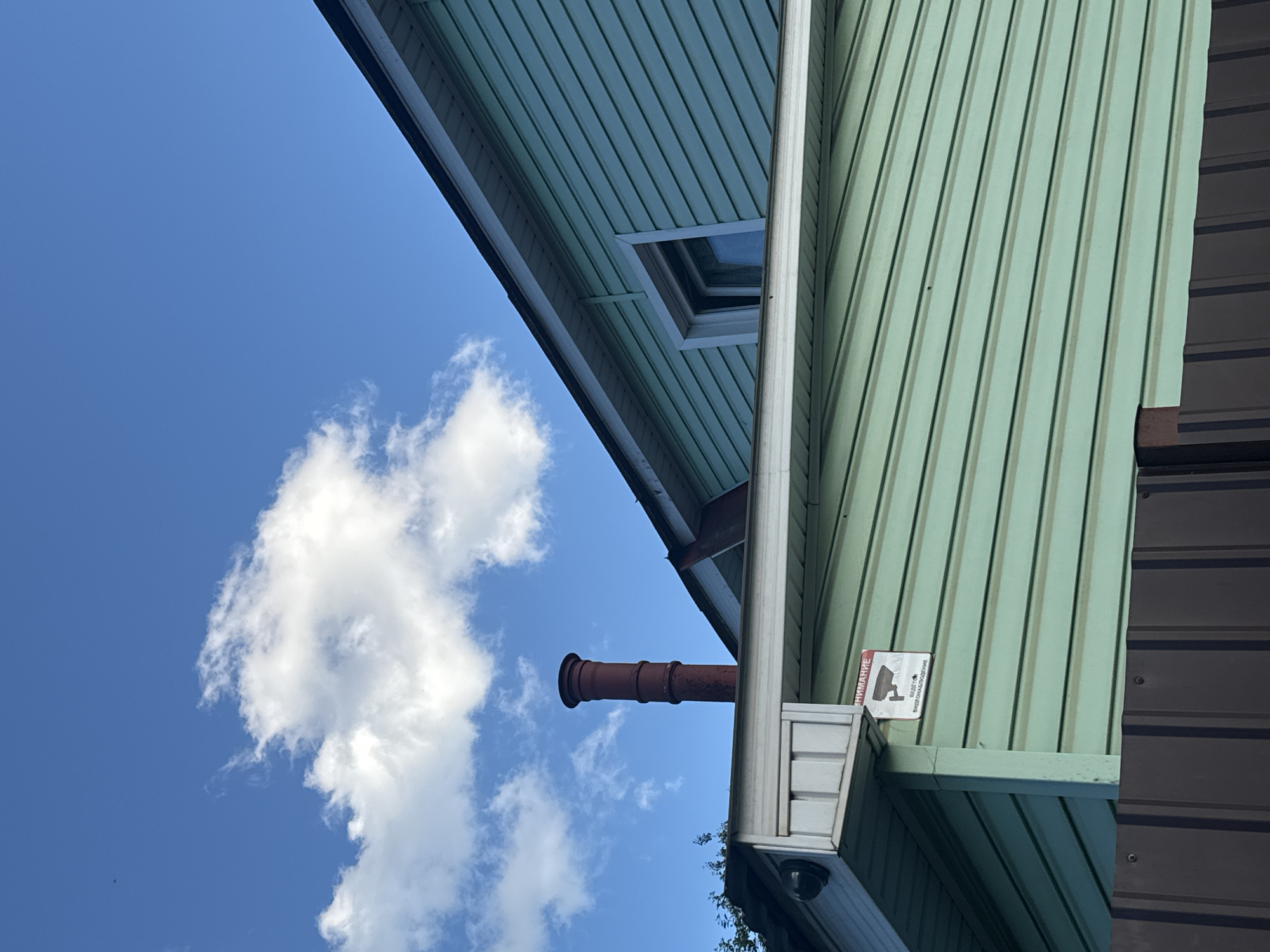The image shows the side and roof of a light green, horizontally-sided house under a bright blue sky with a few white clouds. A dark red brick chimney rises from the roof. A black security camera is visible on the left side of the house, near the eaves. A red and white warning sign with an image of a security camera and text in Russian indicating "ATTENTION: VIDEO SURVEILLANCE" is attached to the side of the house. A dark brown corrugated metal fence runs along the bottom of the image. The scene appears to be an exterior view of a residential building.