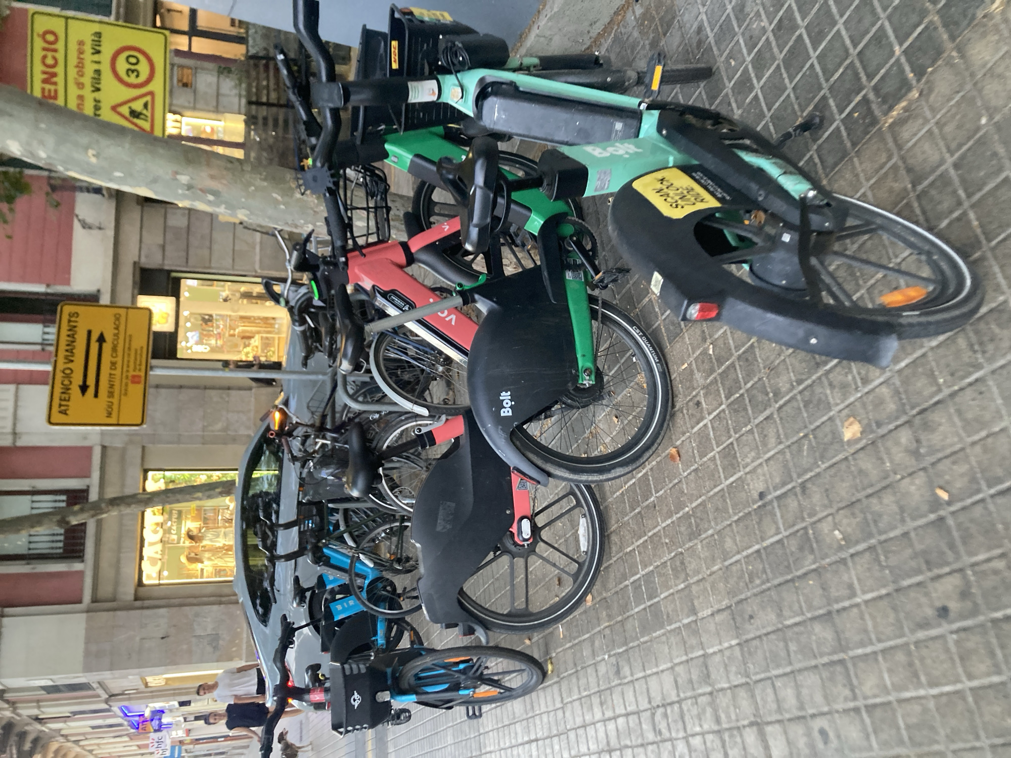 The image depicts a street scene in Barcelona, Spain, likely during the daytime. In the foreground, several shared electric bicycles, identified by the "Bolt" branding, are parked on a tiled sidewalk. One green bicycle is prominently in focus, displaying a "SCAN UNLOCK RIDE" sticker. Other bicycles, in blue and red, are visible behind it. A silver car is parked on the street to the left, with two individuals, a man and a woman, walking past it, accompanied by a small dog. The background shows a building with shopfronts and signage, including an "ATENCIO VIARANTS" (Pedestrian Attention) sign with an arrow indicating two directions. Another yellow sign with a construction symbol and "30" is also visible, suggesting a speed limit or road closure. The environment is urban, characterized by buildings, a sidewalk, street, and parked vehicles.