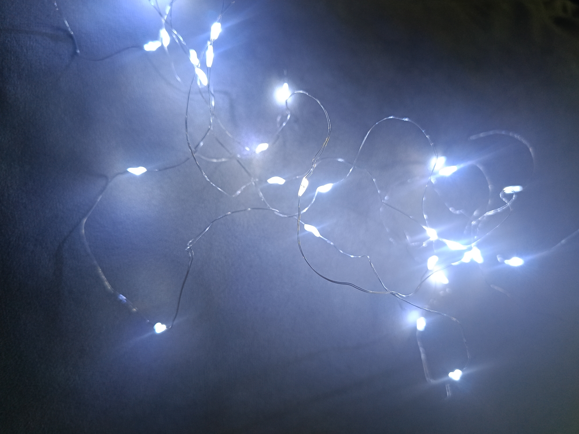 The image captures a string of small, luminous, cool-white LED fairy lights against a dark, textured background. The lights are intricately arranged on a surface that appears to be soft fabric or textile, possibly a blanket or upholstery, displaying subtle undulations and shadows. Each individual light source emits a bright, intense glow, creating significant lens flare and a soft, ethereal halo that illuminates the surrounding dark area with a bluish tint. The thin, metallic wires connecting the LED bulbs are visible, snaking across the frame.
The overall scene suggests an indoor setting, likely in a bedroom or living space, in Chaikovsky, Russia, during nighttime or in a dimly lit environment, given the striking contrast between the intense light and the dark background. The lights seem to be used for decorative purposes or to provide ambient illumination, creating a cozy or atmospheric mood. No specific activity or event is taking place, and no people or visible text are present in the image.