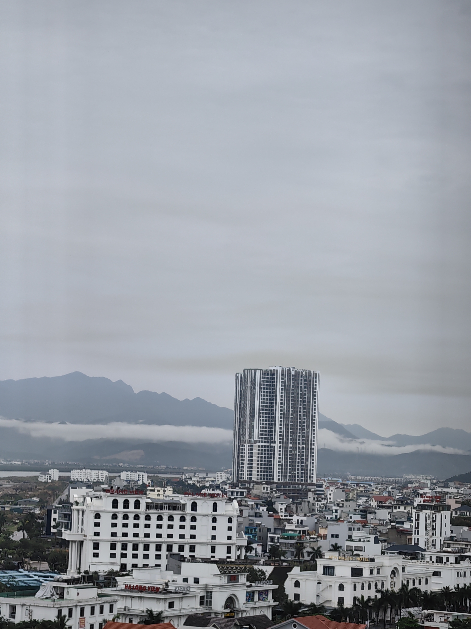 The image shows an elevated view of Hong Gai Ward, Vietnam on an overcast day. A modern, high-rise apartment building dominates the skyline, set against a backdrop of misty, mountainous terrain. In the foreground, a mix of low-rise buildings with white facades and red-tiled roofs fill the area below, with some businesses identified by signage. The sky is uniformly gray, suggesting a cloudy or foggy atmosphere. No people are visible.