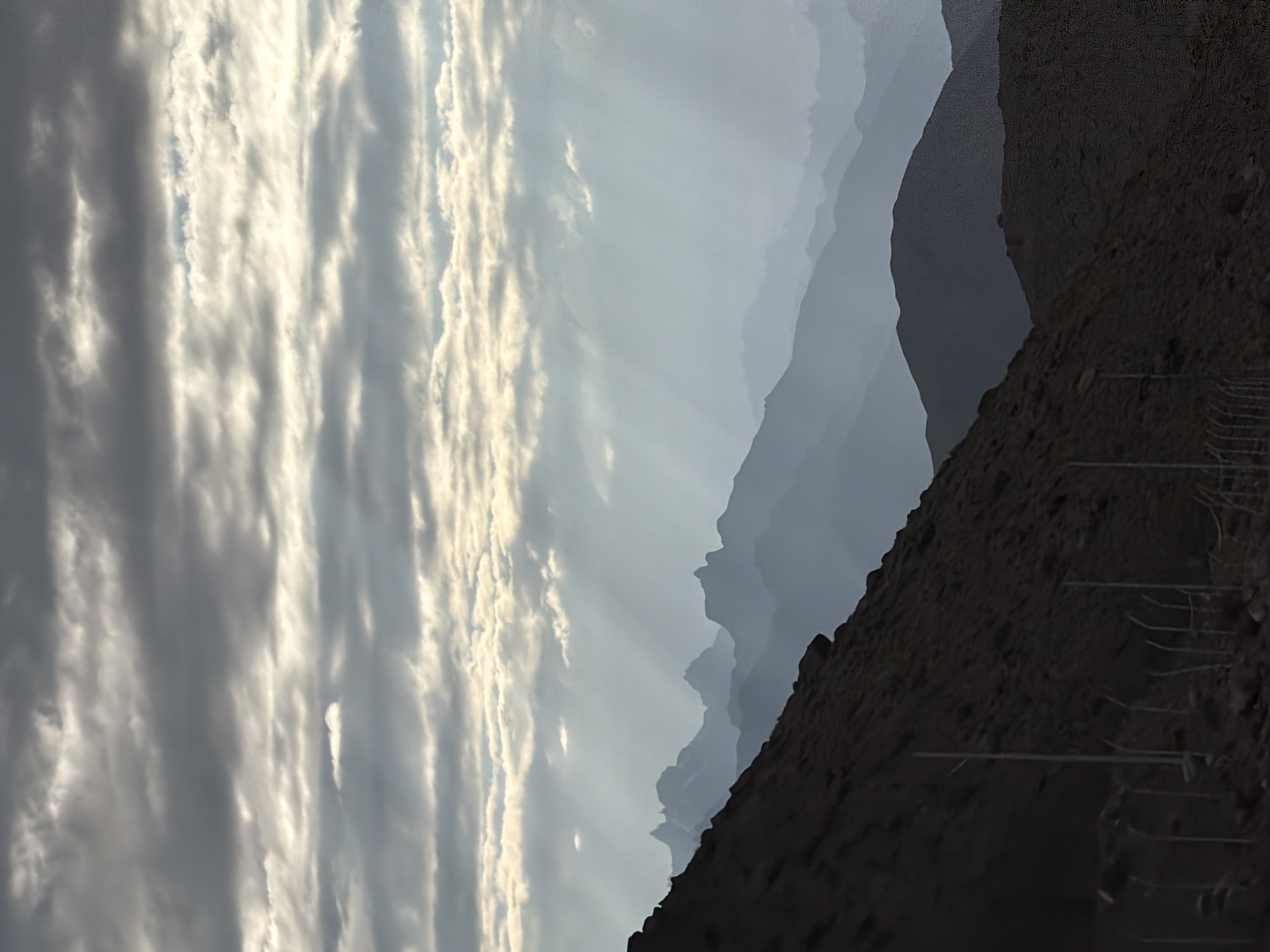 The image depicts a mountainous landscape under a cloudy sky. Sunbeams are visible breaking through the clouds, casting rays onto the distant mountain ranges which fade into a hazy blue silhouette. In the foreground, a dark, rocky slope dominates the lower left of the frame. A fence with posts is visible running horizontally across the bottom portion of this slope. The overall scene conveys a sense of natural grandeur and atmospheric conditions. The provided information about the location in Chumkar, India, is not visually verifiable from the image itself.