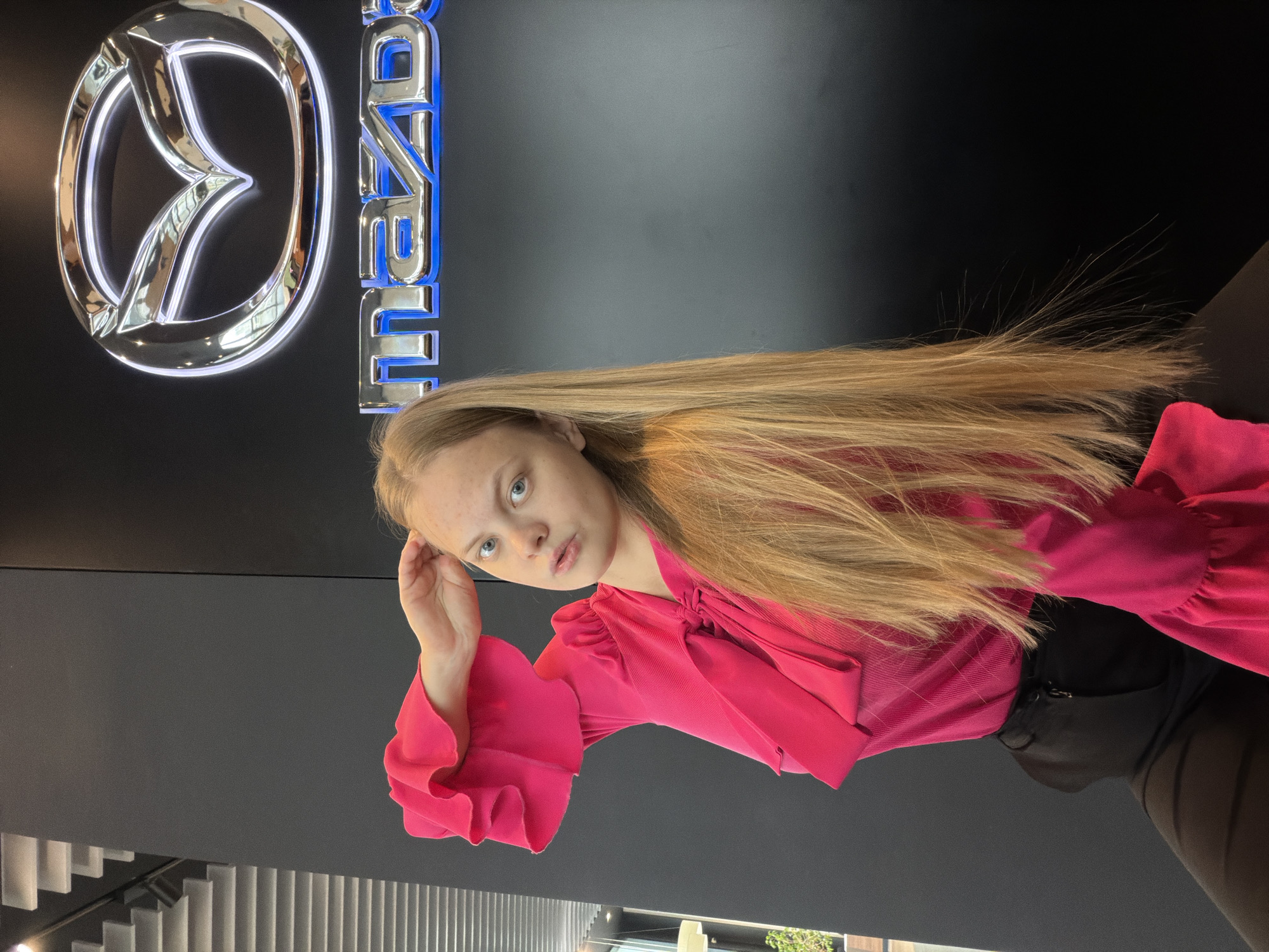 A young woman stands indoors, posing for a photo in front of a Mazda logo. The logo, consisting of the Mazda emblem and the brand name, is illuminated with a blue outline and silver fill. The woman is wearing a bright pink blouse with ruffled sleeves and a tied neck, and black pants or tights. Her long, light brown hair is draped over her shoulder. The background is dark and appears to be a wall or partition. The location is likely a Mazda dealership, although the city of Izhevsk, Russia, cannot be confirmed without further information. A staircase with white vertical elements is visible in the bottom left corner.