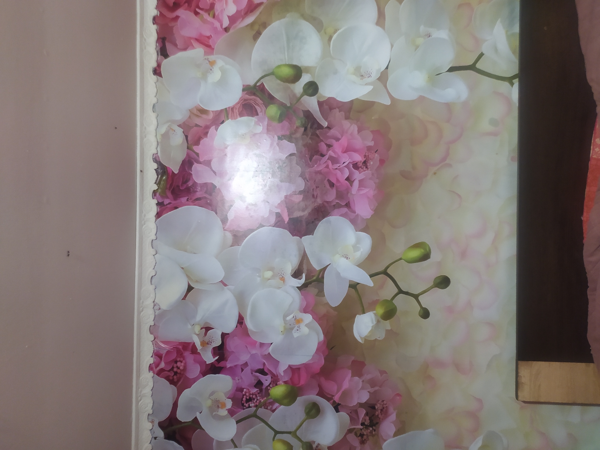 The image shows a close-up view of a wall adorned with a floral wallpaper featuring white orchids and pink flowers. The wallpaper has a textured appearance and is detailed, with individual petals and buds clearly visible. The lighting in the image suggests an indoor setting, possibly a bedroom or living area. The top of the image shows a portion of a ceiling and a decorative trim. The bottom of the image reveals the top of a dark brown headboard of a bed, with some bedding visible underneath. The overall scene is static and does not depict any people or actions. There is no information in the image that indicates a location in the city of Hoverla, Ukraine.