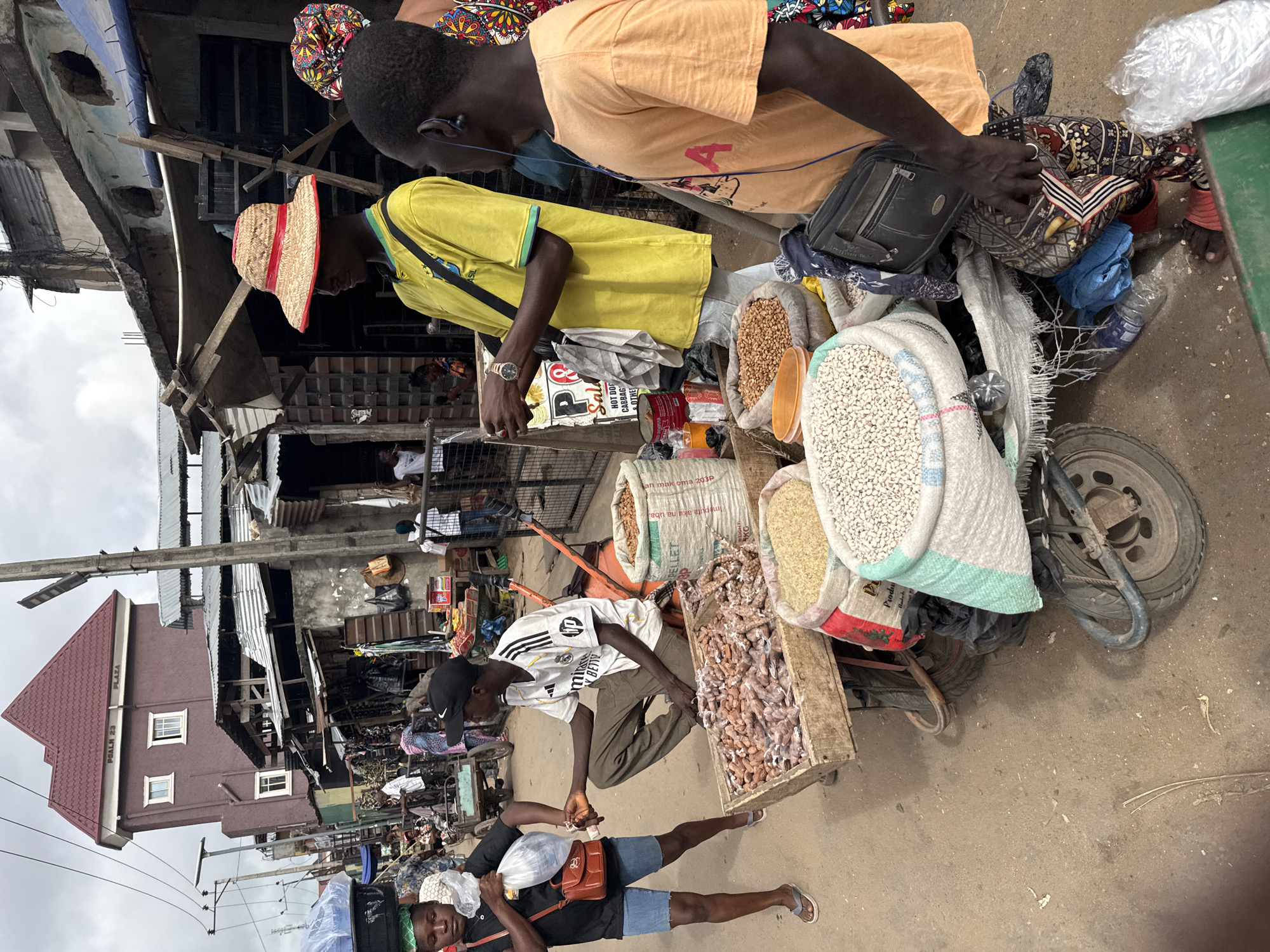 Here's a description of the image:
The image captures a bustling street scene in Onitsha, Nigeria. It's an outdoor setting, with a variety of street vendors and pedestrians navigating a crowded marketplace. Several men are visible, engaged in transactions and selling goods. The vendors are selling various food products including beans, grains, and dried seafood. The goods are displayed in large sacks and wooden crates, loaded onto a cart. The background shows buildings with a mix of materials, including corrugated metal roofs, along with a Plaza building. Power lines are visible in the upper left. The environment suggests a typical busy market day in a Nigerian city.