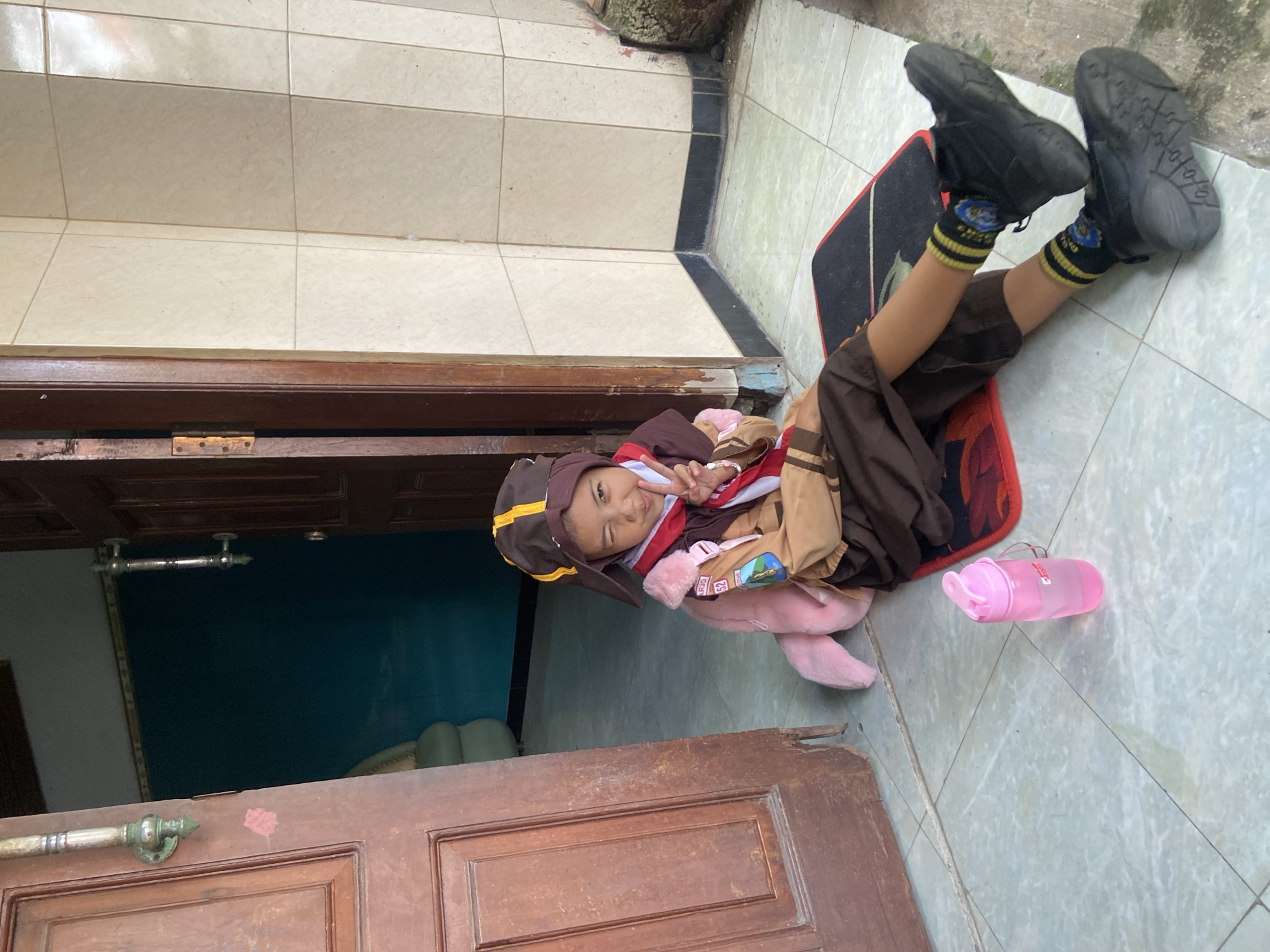 A young person wearing a scout uniform is lying on a rug in front of an open door. The scout uniform consists of a brown shirt, dark brown shorts, a brown hat with a yellow stripe, and black shoes with yellow-striped socks. The person is holding a pink stuffed animal and making a peace sign with their fingers. A pink water bottle is next to the rug. The location appears to be the city of Gelam, Indonesia. The environment consists of tiled flooring and an open door leading into a room with a blue wall.