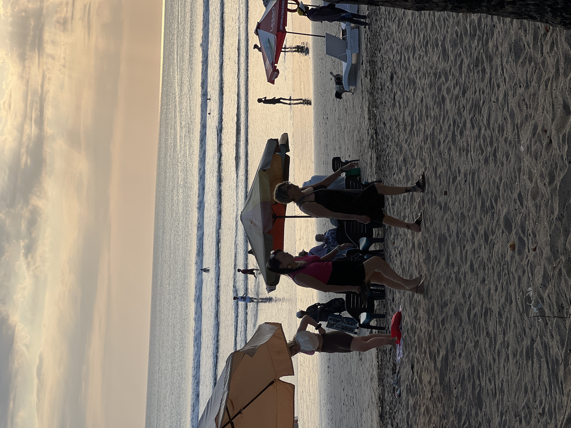 This is a photograph of Kuta Beach in Kuta, Indonesia, taken during the late afternoon or early evening. The sky is a soft gradient of pale orange and yellow, with streaks of grey clouds, indicating either sunrise or sunset. The ocean is calm with gentle waves rolling towards the shore.
In the foreground, on the sandy beach, several people are visible. Two women are walking towards the right of the frame. One is wearing a pink tank top and a black skirt, while the other is in a black dress. Further to the left, a woman with blonde hair is adjusting her white hat, wearing a pink bikini top and brown shorts. Behind them, there are beach chairs and umbrellas, with a few other individuals and a dog in the middle ground. In the ocean, several people can be seen swimming or standing in the shallow water. The overall atmosphere is relaxed and leisurely, typical of a beach scene.