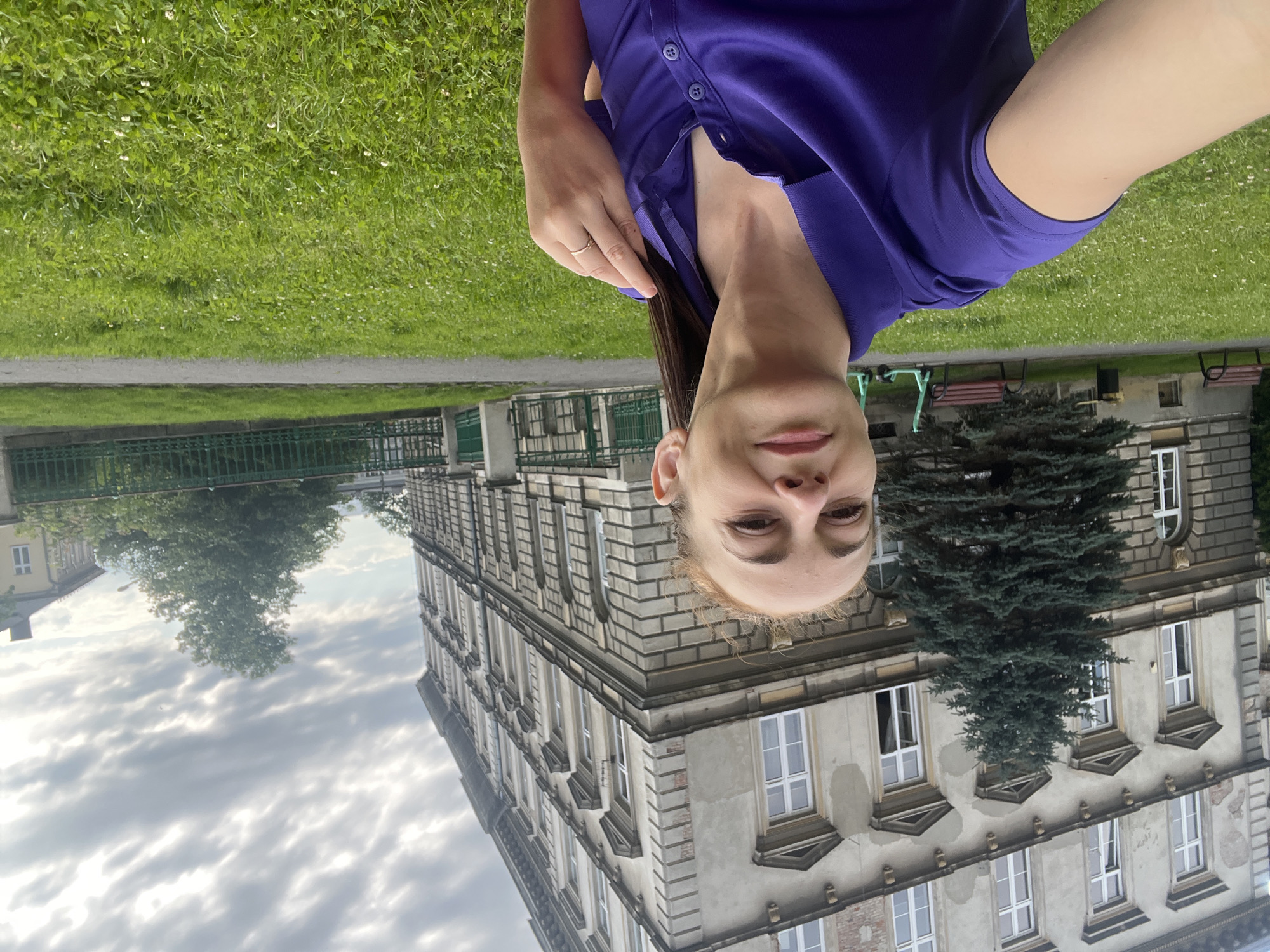 A woman is taking a selfie in front of a large, ornate building. The building appears to be made of stone and has many windows with white frames. In front of the building is a manicured lawn with a large evergreen tree. To the right of the tree are two red benches and a small trash can. Further in the background, there are two green electric scooters and a green metal fence. The sky is overcast. The woman is wearing a purple polo shirt and has her hair pulled back in a ponytail. She is smiling and touching her hair with her right hand. The image is taken from a low angle.