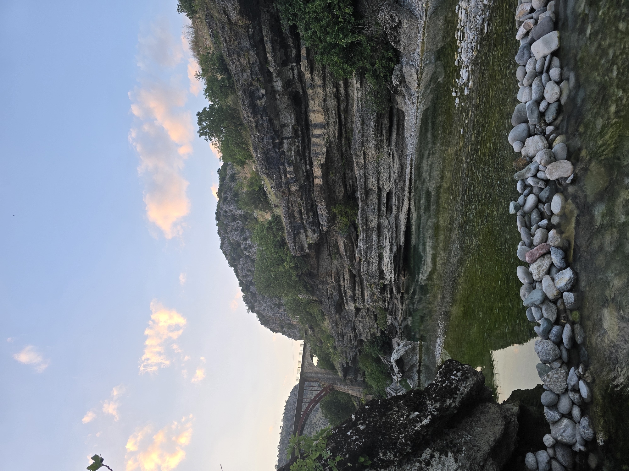 The image depicts a scenic view of the Morača River canyon near Bioče, Montenegro. In the foreground, a line of smooth, grey and pink river stones forms a natural border along the shallow water's edge. The river itself is clear, with visible pebbles on the riverbed and a greenish hue. In the midground, the canyon walls rise steeply on either side, characterized by layered rock formations and patches of green vegetation. A large, dark rock with moss is prominent on the left. Further in the distance, an arched bridge, with a red structure visible underneath, spans the canyon, suggesting it is a road or railway bridge. The sky above is a soft blue with scattered, pale pink clouds, indicative of either dawn or dusk. The overall impression is one of natural beauty and tranquility.
