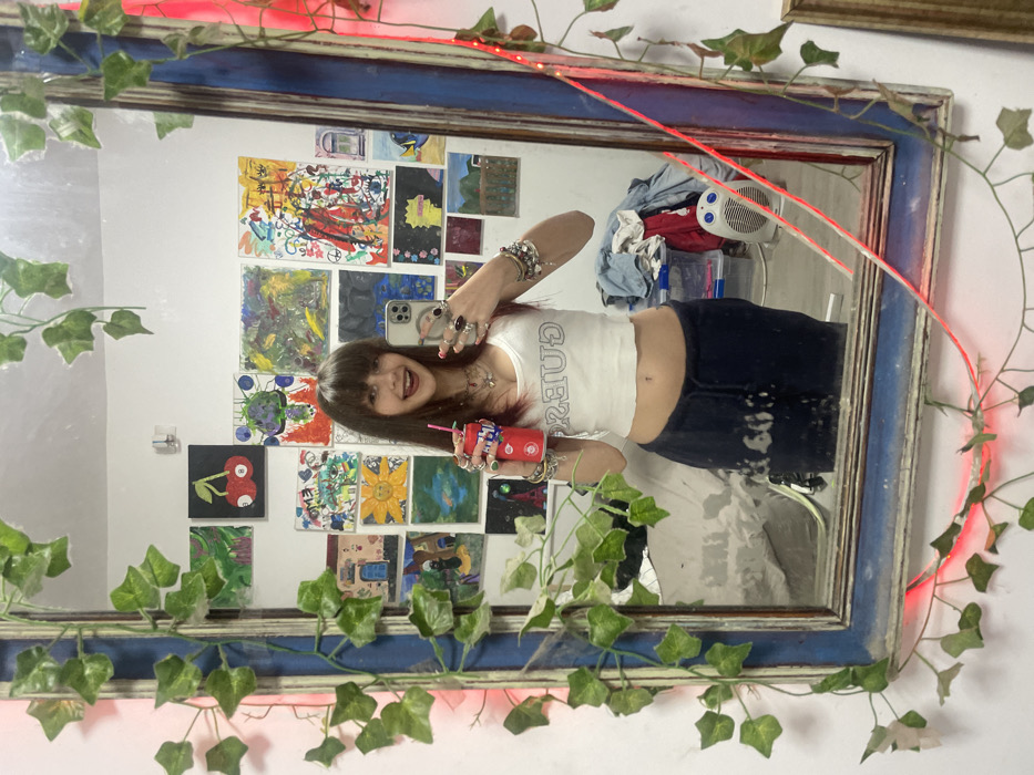A young woman is captured in a mirror selfie, grinning broadly. The image is oriented vertically, showing her from the waist up. She has long, straight dark hair with bangs and a wide, open-mouthed smile, revealing her teeth. She wears a white cropped tank top with the word "GUESS" printed in gray capital letters across the chest, exposing her midriff and navel. Her lower body is clad in dark blue or black low-rise bottoms.
In her left hand (viewer's right), she holds a smartphone, likely taking the photo. This hand is adorned with multiple rings on her fingers. Her right hand (viewer's left) holds a red plastic cup with a pink straw, decorated with several colorful bracelets on her wrist. Her fingernails appear to be painted a dark color.
The reflection in the mirror reveals a white wall behind her, extensively decorated with an array of small to medium-sized artworks, primarily colorful paintings or drawings. These artworks are varied in style and content, suggesting a collection of personal pieces or art. Notable pieces include an abstract red painting, a black canvas with two red cherries, and a yellow artwork resembling a sun.
To the right of her reflection (viewer's left), a white fan or heater unit with a blue dial is visible, along with some loosely folded blue and red clothing. Below her reflection, a portion of a white-sheeted bed or furniture is discernible.
The mirror itself has a distressed frame, predominantly blue with hints of a lighter, possibly gold or brown, undertone. Green artificial ivy vines are draped decoratively around the top and sides of the mirror frame, extending onto the surrounding white wall. A thin red LED light strip is also wound around the mirror frame, casting a subtle red glow. The overall setting appears to be an interior, likely a personal room.