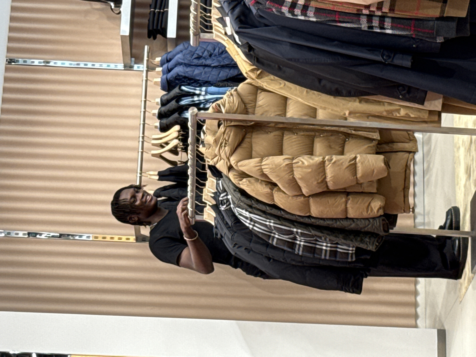 In a retail clothing store in La Roca del Vallès, Spain, a young Black man with dreadlocks is seen browsing a rack of puffer jackets. He is wearing a black t-shirt and black pants. The store features beige, pleated curtains as a backdrop and displays a variety of outerwear including puffer jackets in brown and blue, as well as plaid jackets. The man is looking at the clothes and appears engaged with the selection.