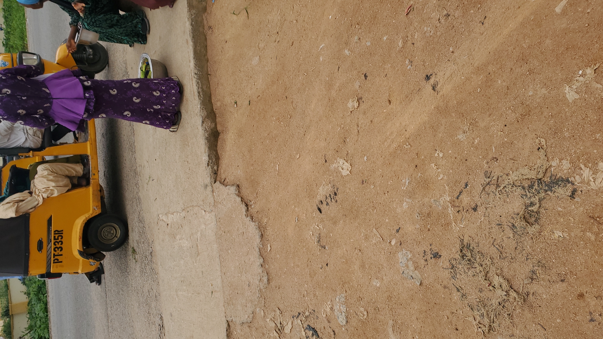The image depicts a street scene in Jalingo, Nigeria, with a yellow motorized tricycle, commonly known as an "achaba" or "keke," parked on the side of a paved road. The tricycle displays the license plate "PT-335R." A person wearing a purple patterned dress with a ruffled peplum is standing to the right of the tricycle, facing away from the camera. Another person, partially visible, is seated inside the tricycle. The ground is a mix of dirt and concrete, with debris scattered across the surface. A metal bowl containing what appears to be green produce is placed on the ground near the standing person. In the background, vegetation and a yellow structure are visible, suggesting an urban or semi-urban environment.