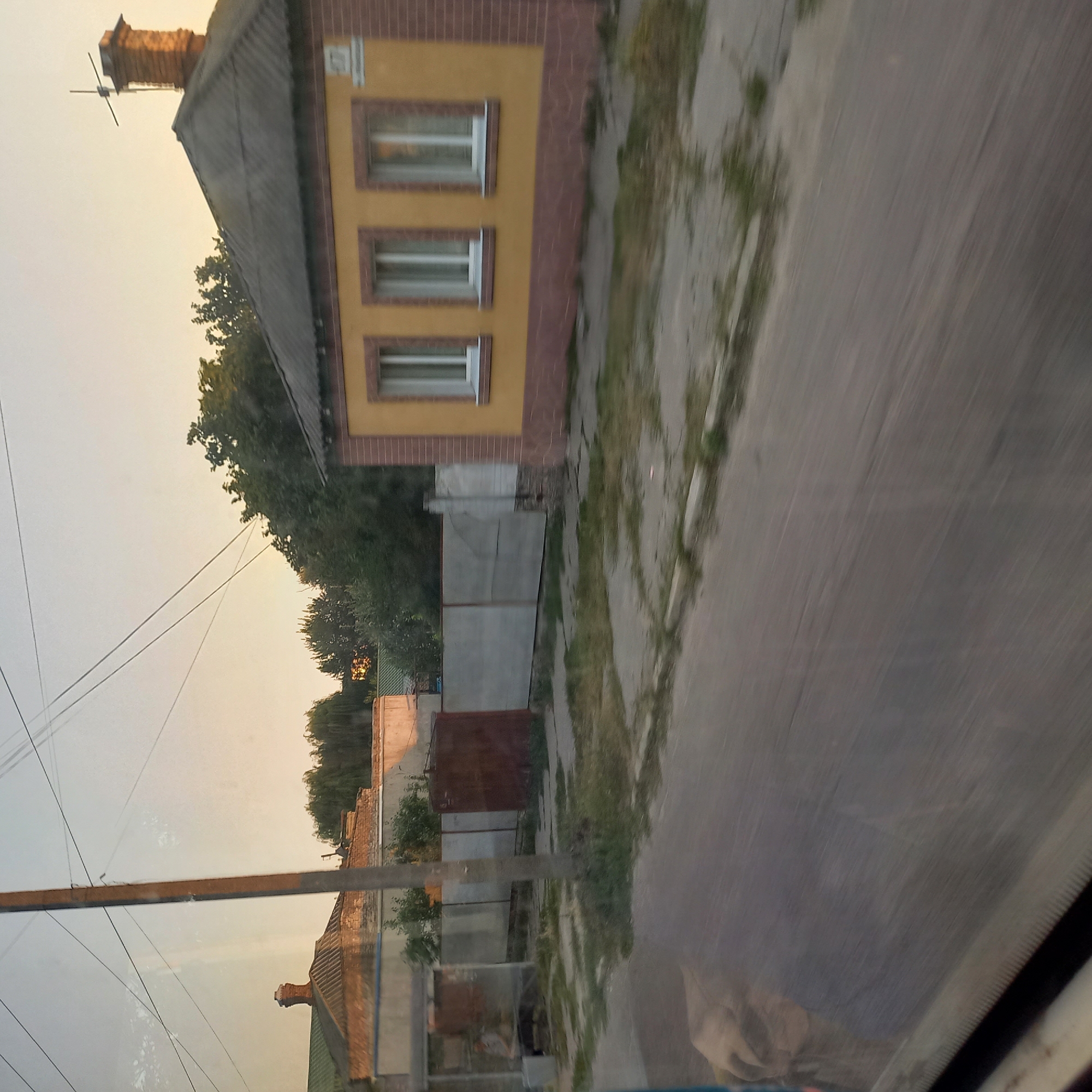 The image depicts a street scene in Kropyvnytskyi, Ukraine, likely during the day. The foreground is dominated by a blurred asphalt road, indicating movement. In the midground, a yellow building with a brown-tiled base and a gray roof is visible. It features three white-framed windows and a brick chimney with an antenna on top. Adjacent to this building is a concrete fence with a rusty metal gate. To the left, a bus stop structure with a person sitting inside is visible, along with another rusty metal structure. The background consists of more buildings, trees, and overhead power lines against a pale sky. The overall impression is one of a residential or commercial area in a Ukrainian city.