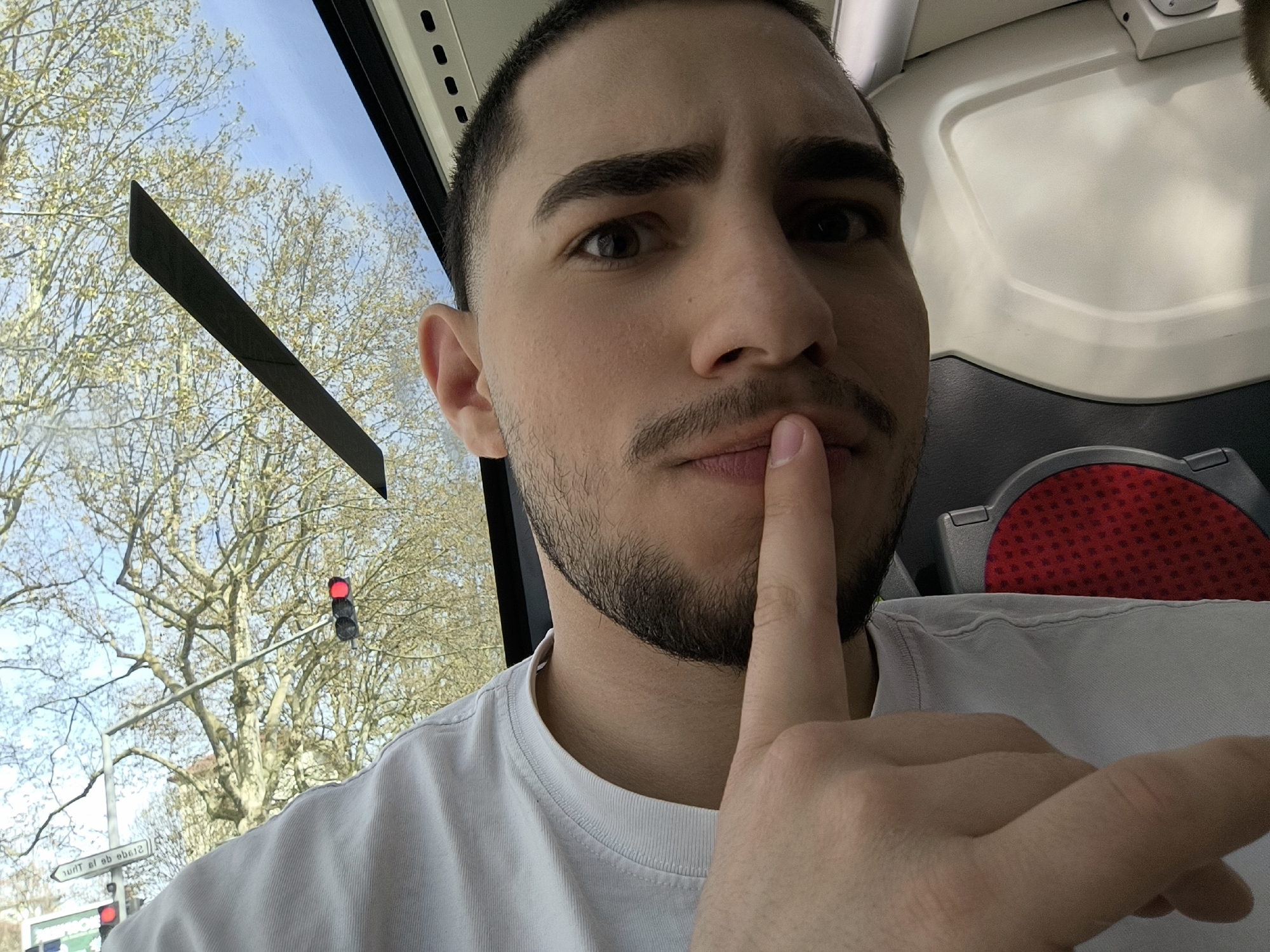 A medium-shot selfie of a man with dark hair and a beard is taken from the interior of a moving bus. The man is Caucasian, wearing a white t-shirt, and holding his index finger to his lips in a gesture of silence.
Through the bus window, a street scene is visible. A red traffic light is mounted on a pole, with bare trees filling the background under a blue sky. A street sign indicates the location is Strasbourg, France.
