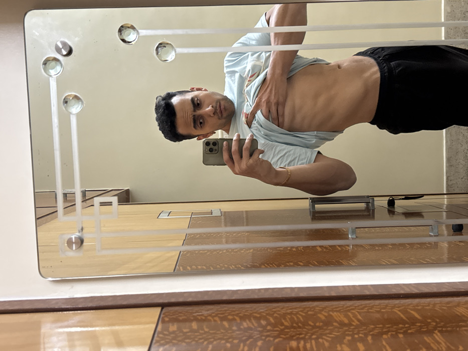 A man takes a mirror selfie, revealing a toned physique. He wears black pants and a light blue t-shirt pulled up, exposing his abdomen. The reflection is in a vintage-style mirror with geometric designs and glass embellishments. The background shows a neutral-toned wall and a partially visible wooden surface. The man holds a phone in his hand, presumably taking the picture.