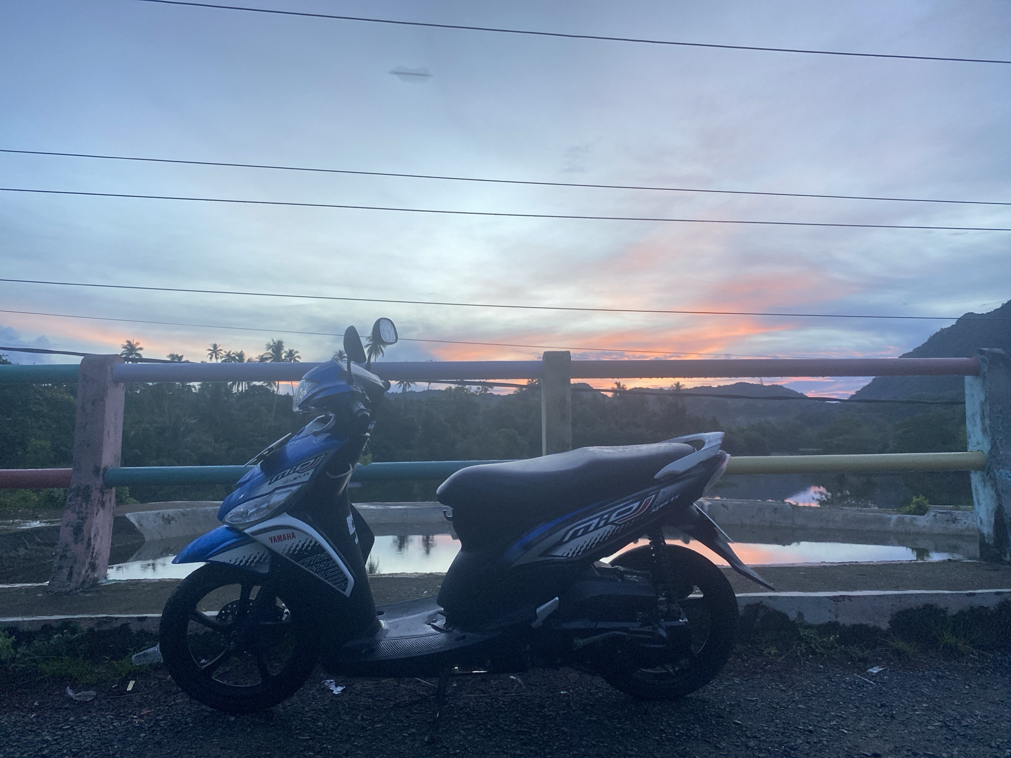 The image depicts a serene twilight scene with a blue and white Yamaha Mio scooter parked on a gravel surface. The scooter is positioned on the right side of the frame, facing left. Beyond the scooter, a low-lying concrete barrier with colorful railings runs horizontally across the mid-ground. A body of water, reflecting the sky, is visible behind the barrier. In the distance, lush green hills and palm trees are silhouetted against a sky filled with pastel hues of pink, orange, and blue, indicative of a sunset or sunrise. The overall atmosphere is tranquil. This scene is located in Lambaro Kueh, Indonesia.