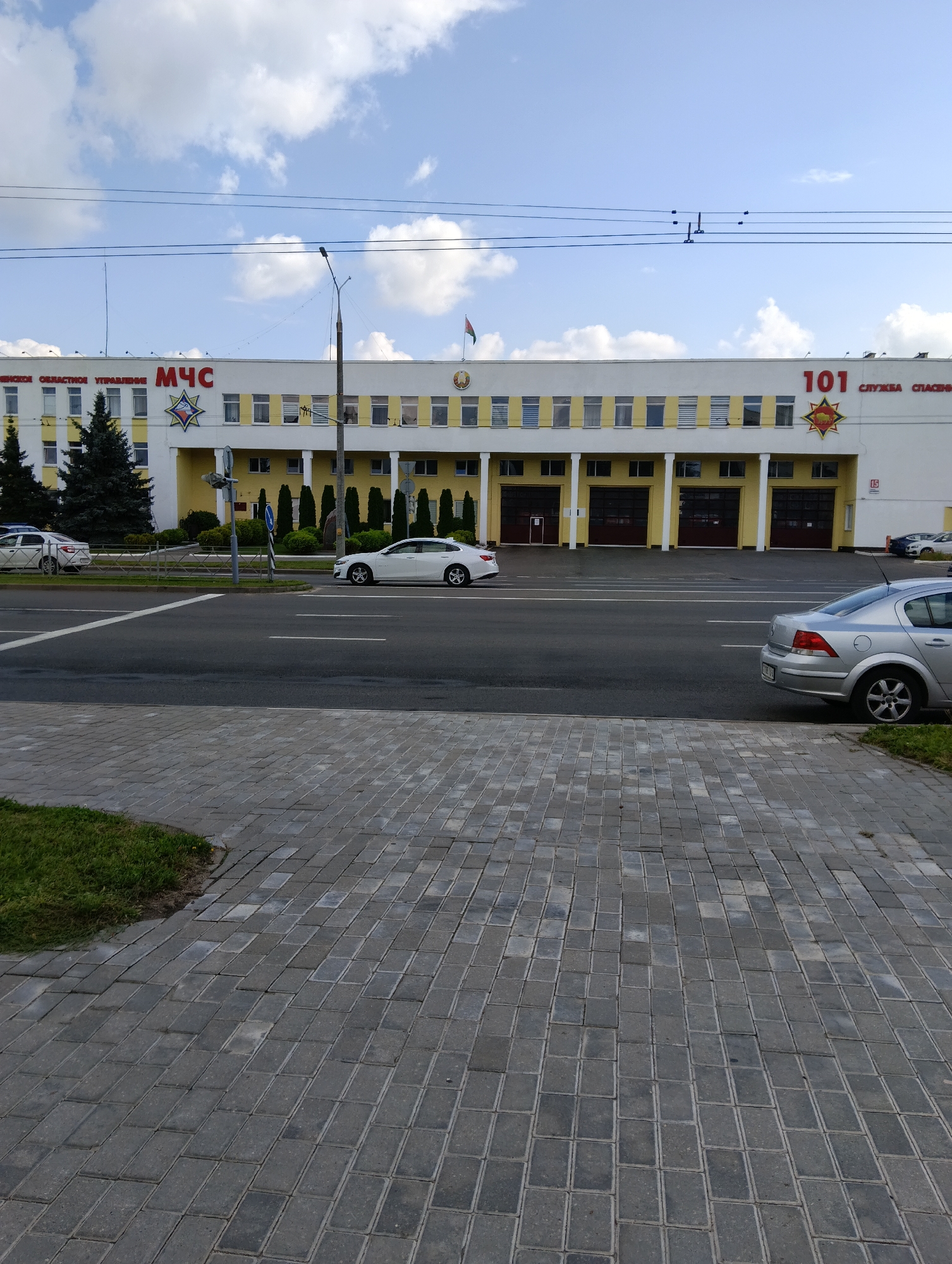 The image shows a wide, paved street in front of a multi-story building. The building appears to be a public service building, with "МЧС" (Ministry of Emergency Situations) and "101 СЛУЖБА СПАСЕНИЯ" (101 Rescue Service) signs visible in red lettering. The building has a white facade with yellow accents around the windows and a row of garage doors on the ground floor. Several cars are parked on the street and in front of the building. The sky is blue with scattered white clouds. The street is paved with asphalt, and a sidewalk made of gray interlocking bricks runs along the side of the road. Green grass and trees are visible in front of the building. The scene is set in the city of Hrodna, Belarus.
